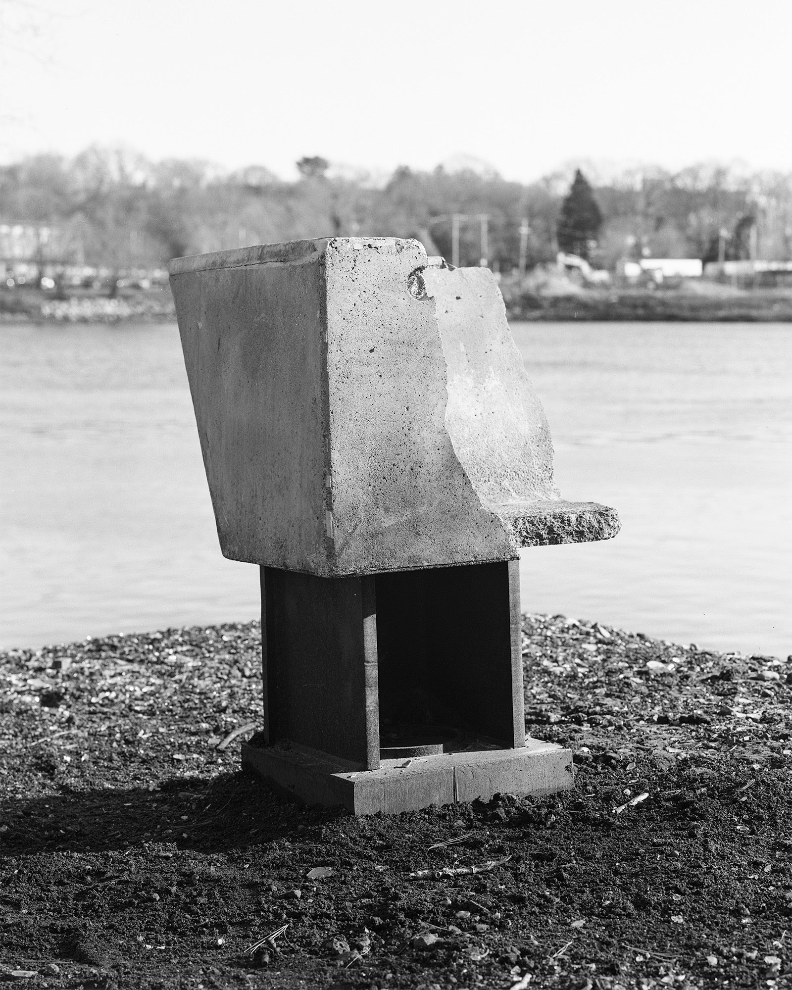 black and white image of concrete sitting atop rusty I-beam metal piece located on the bank of a river