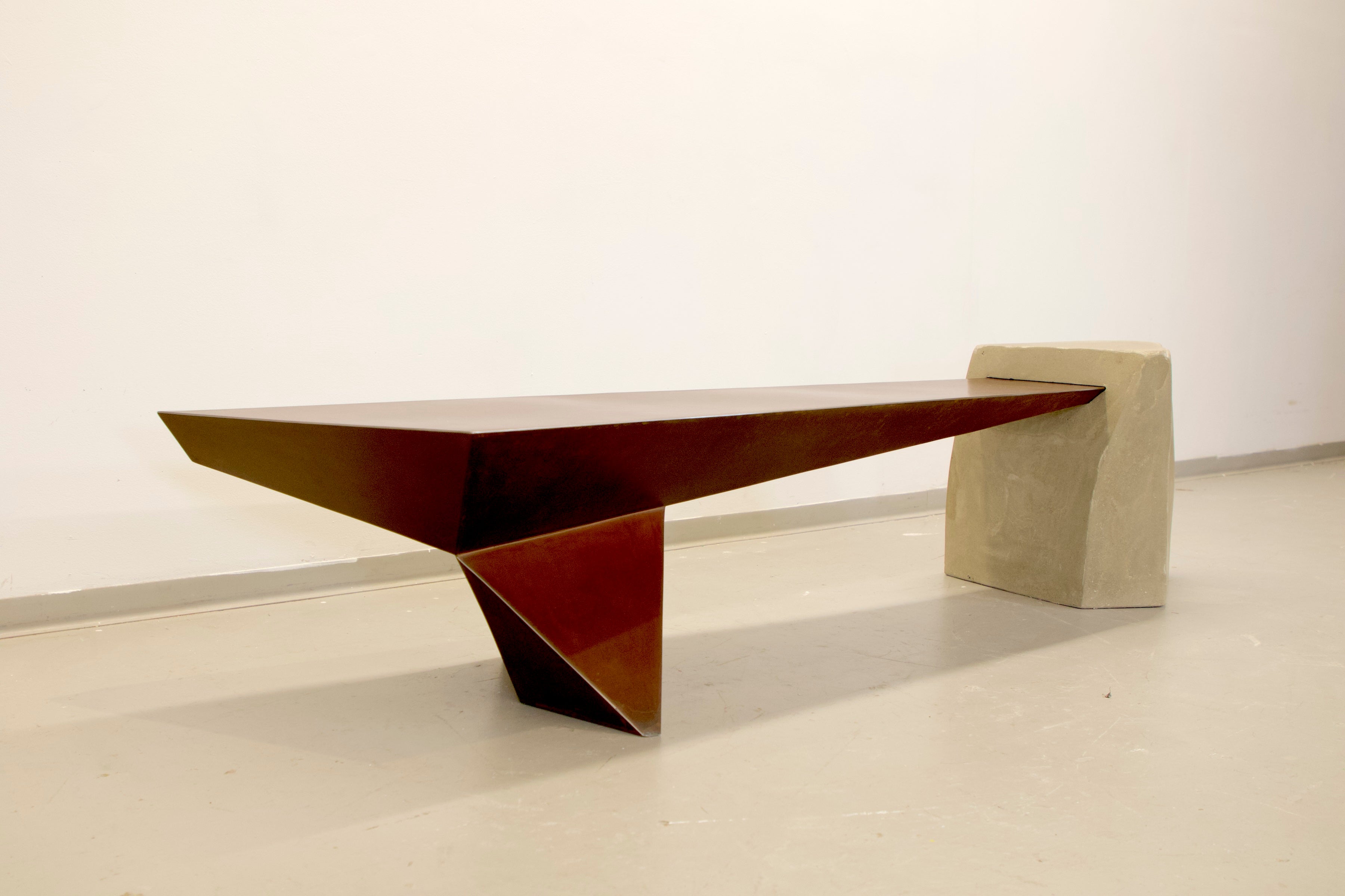 Angular rusty metal bench attached to angular concrete mass, corners of rusty metal have been selectively polished to highlight the edges