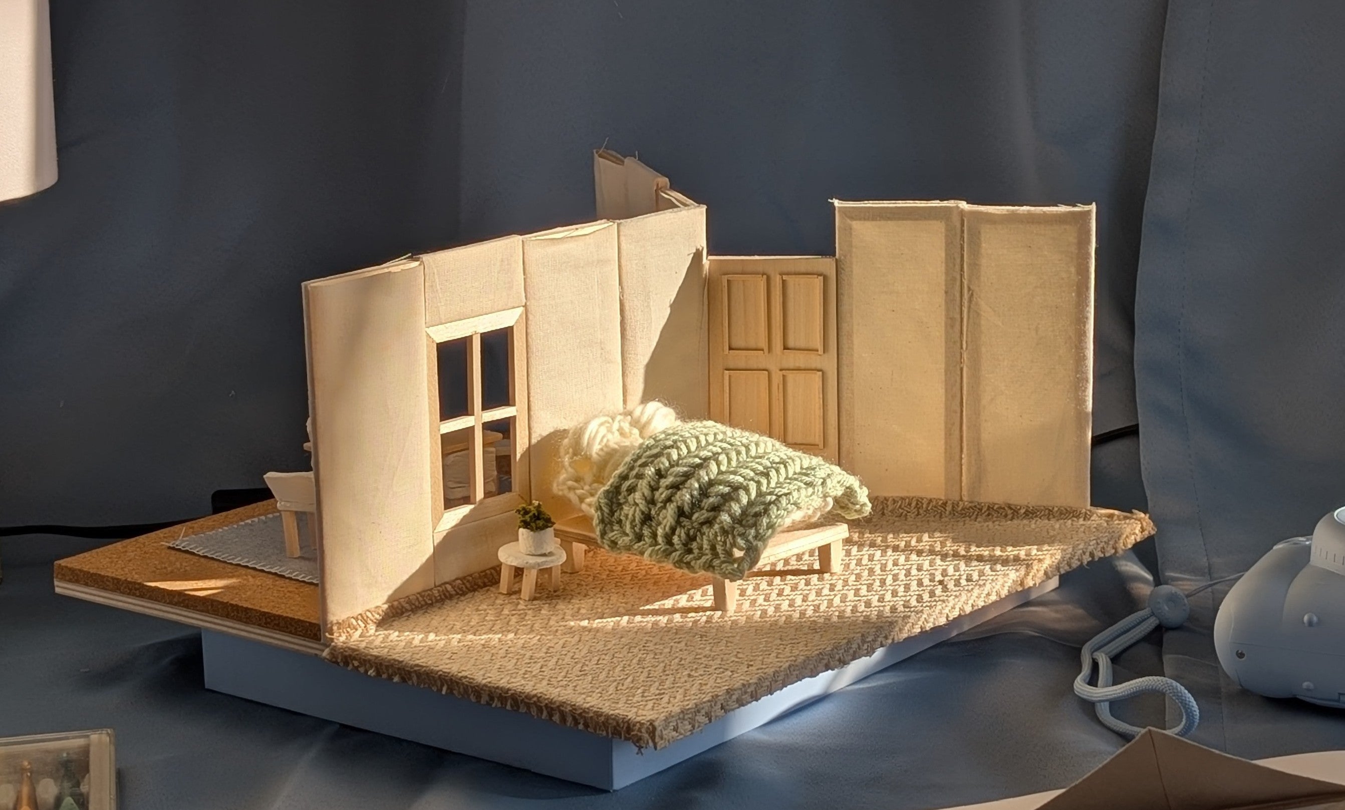 A three way stage model made of textiles, hand knits and crochet. It depicts a bedroom, kitchen, and front porch.