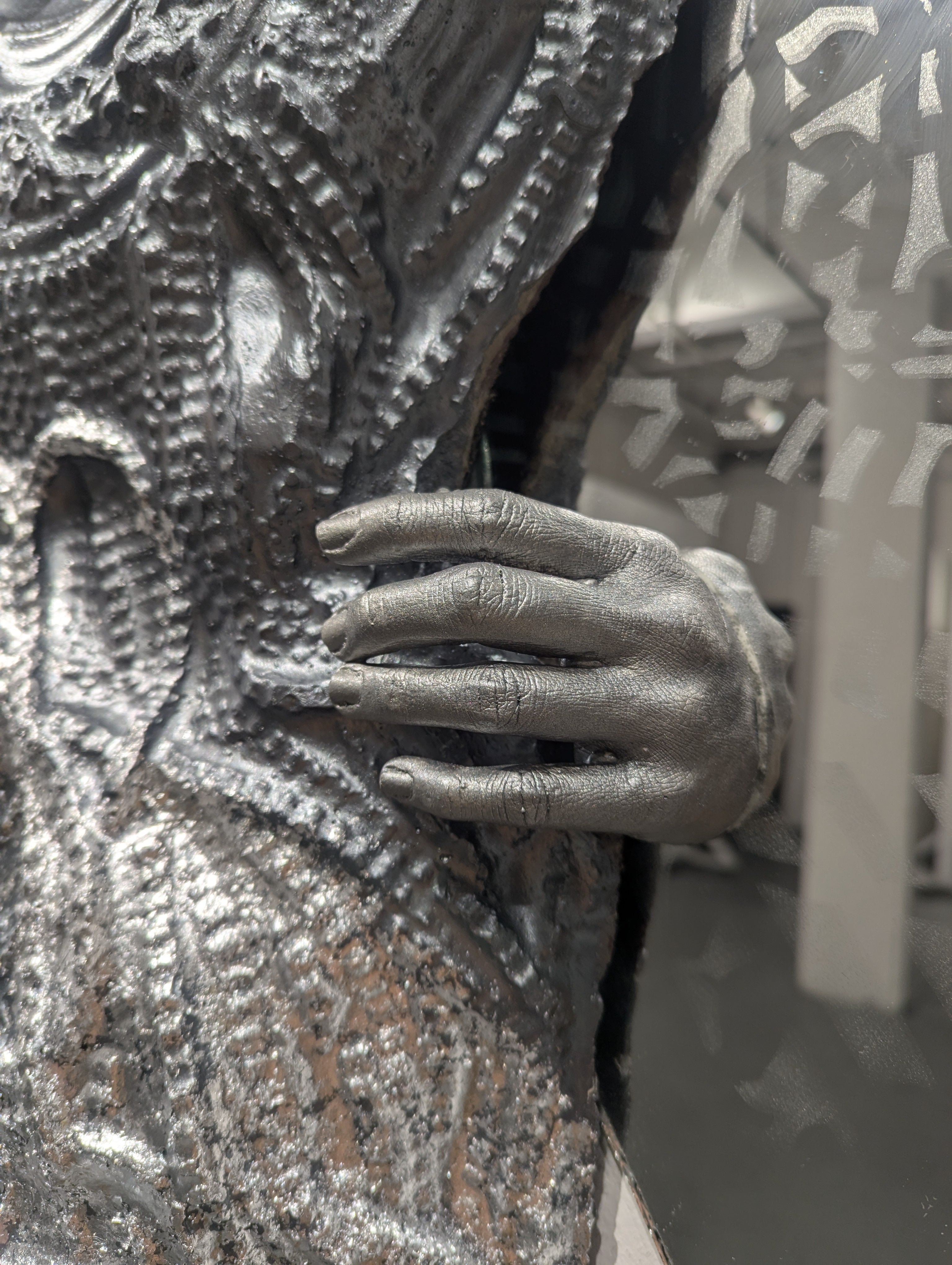 Detail chain-textured cement form, partially silver over gray, surrounded by mirrors, faintly etched with Hmong batik patterns. A silver-gray hand emerges from one mirror, reaching toward the cement in an ambiguous gesture of embrace or pull.