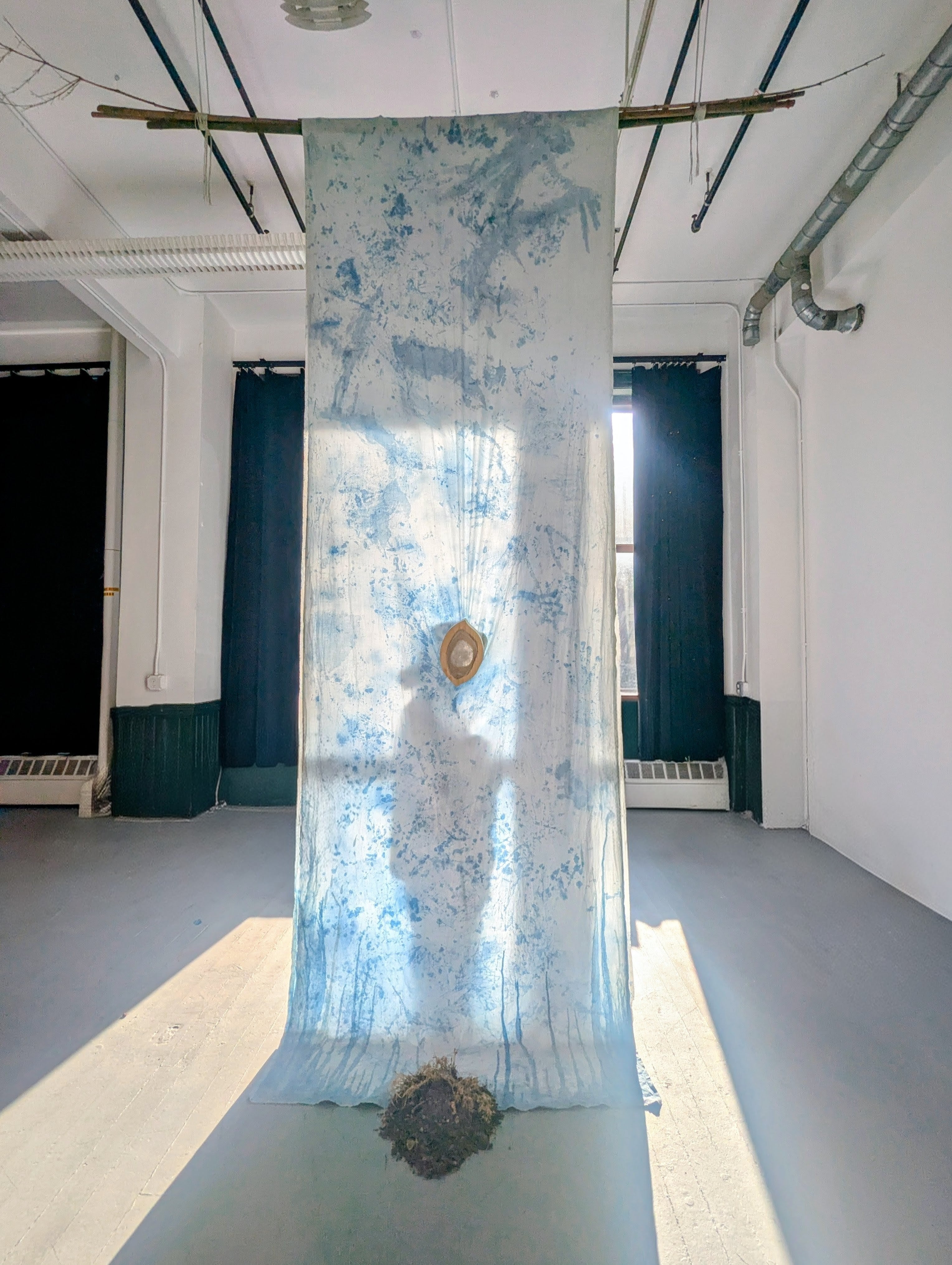 Floor-to-ceiling white cotton draped like a runner, marked with indigo body prints. Backlit to reveal a person's shadow silhouette with a heart-shaped glass above at the center, with dried soil and plants beneath.