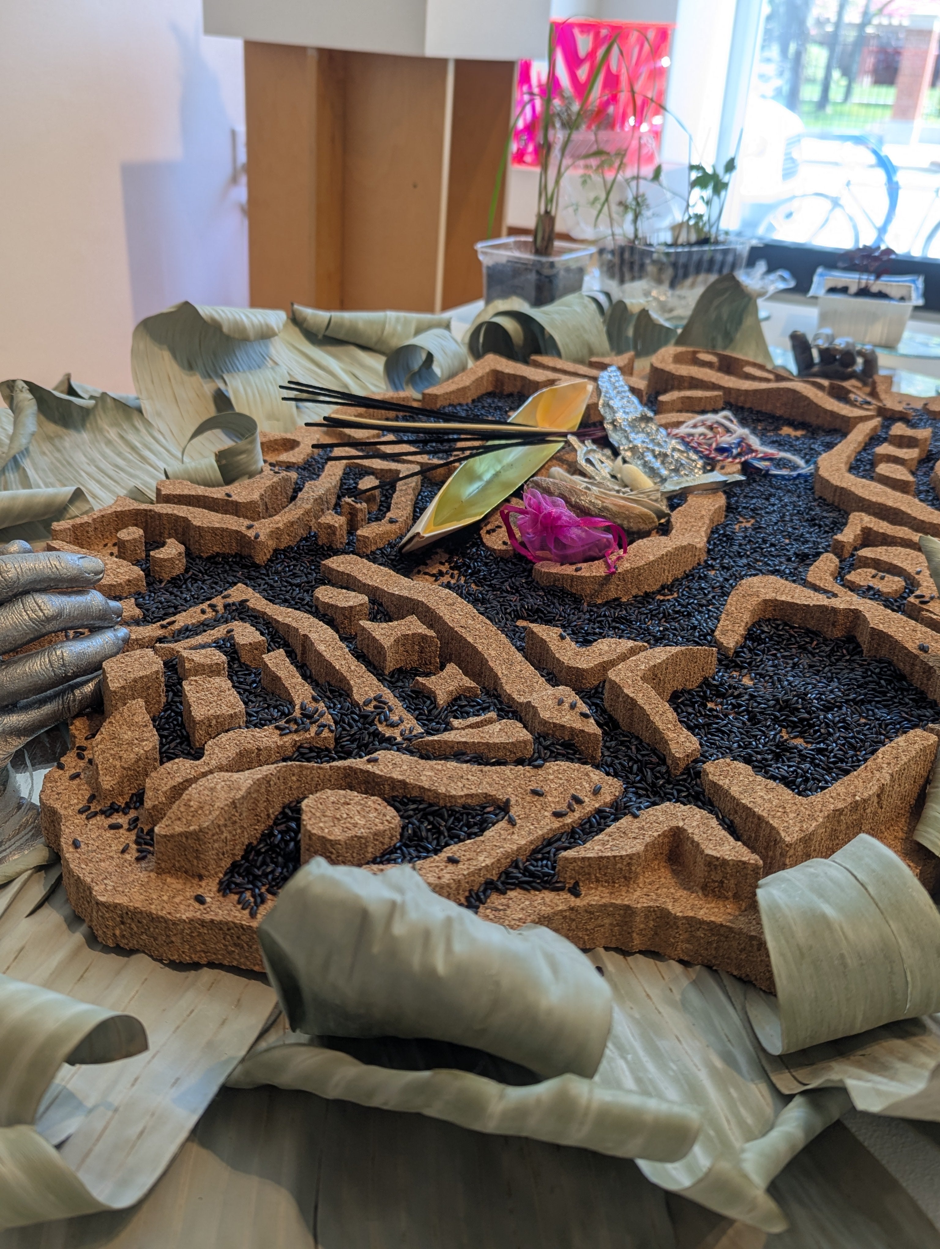 Angled side detail view of Batik-patterned altar covered in black rice and protective talismans, flanked by live herbs and banana leaves.