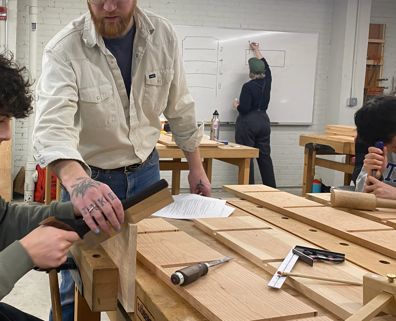 A bearded man holds the saw blade for a teenage boy cutting through a plank of wood in a woodworking classroom.