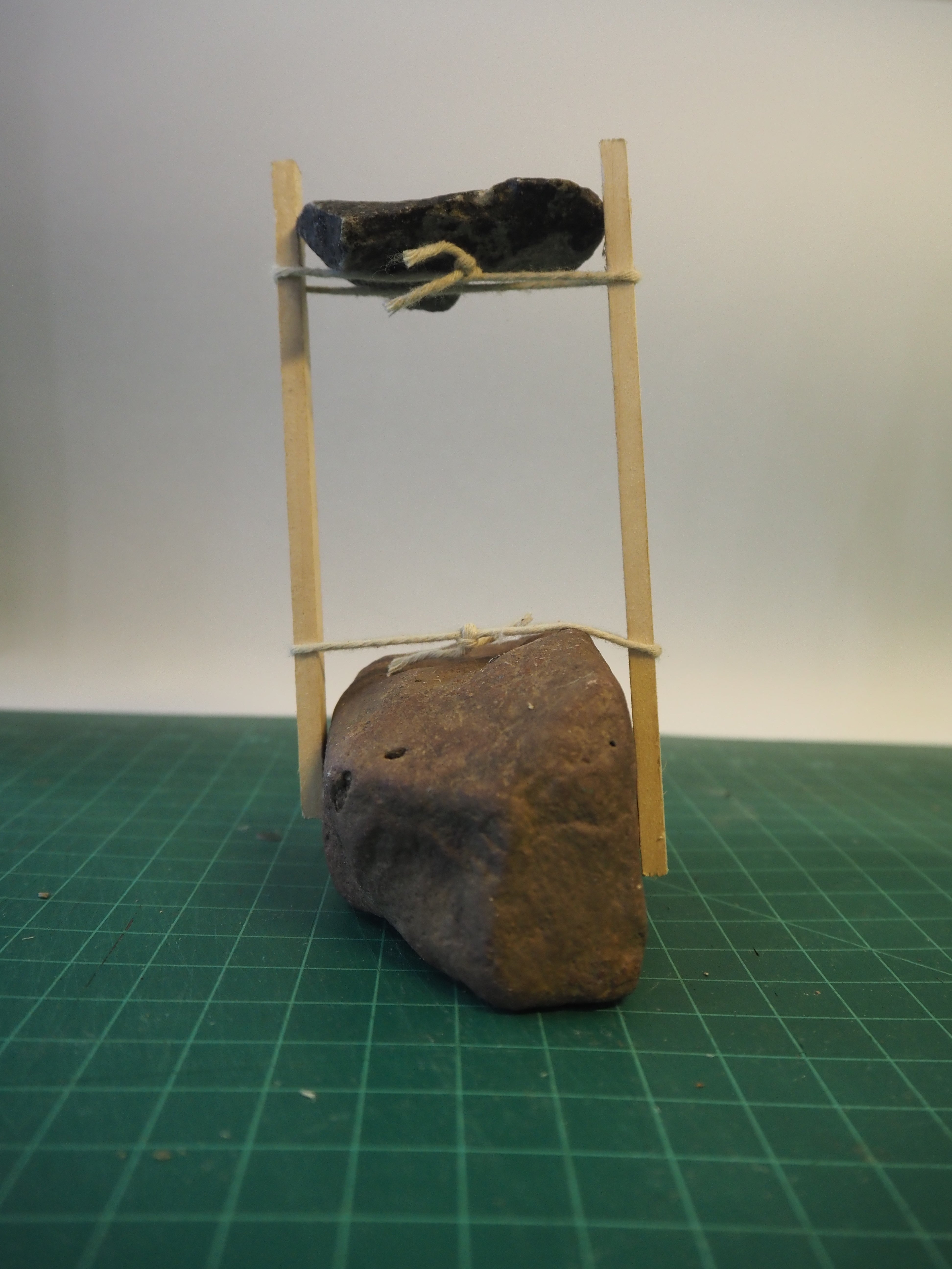 Two wood sticks and string support an elevated rock, anchored by rock on the ground.
