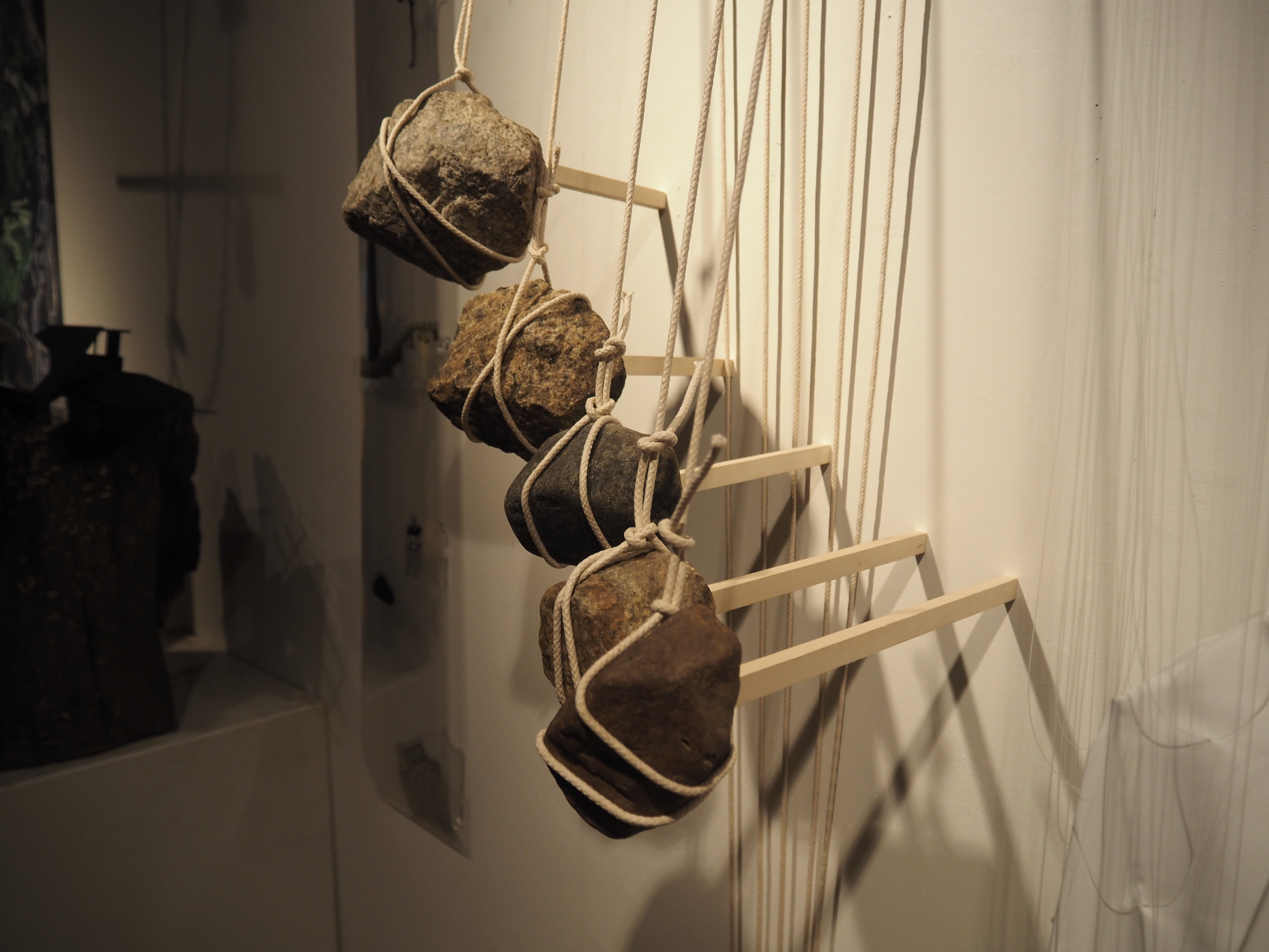 Five rocks suspended by sticks compress five wood sticks against the wall, holding them in the air.