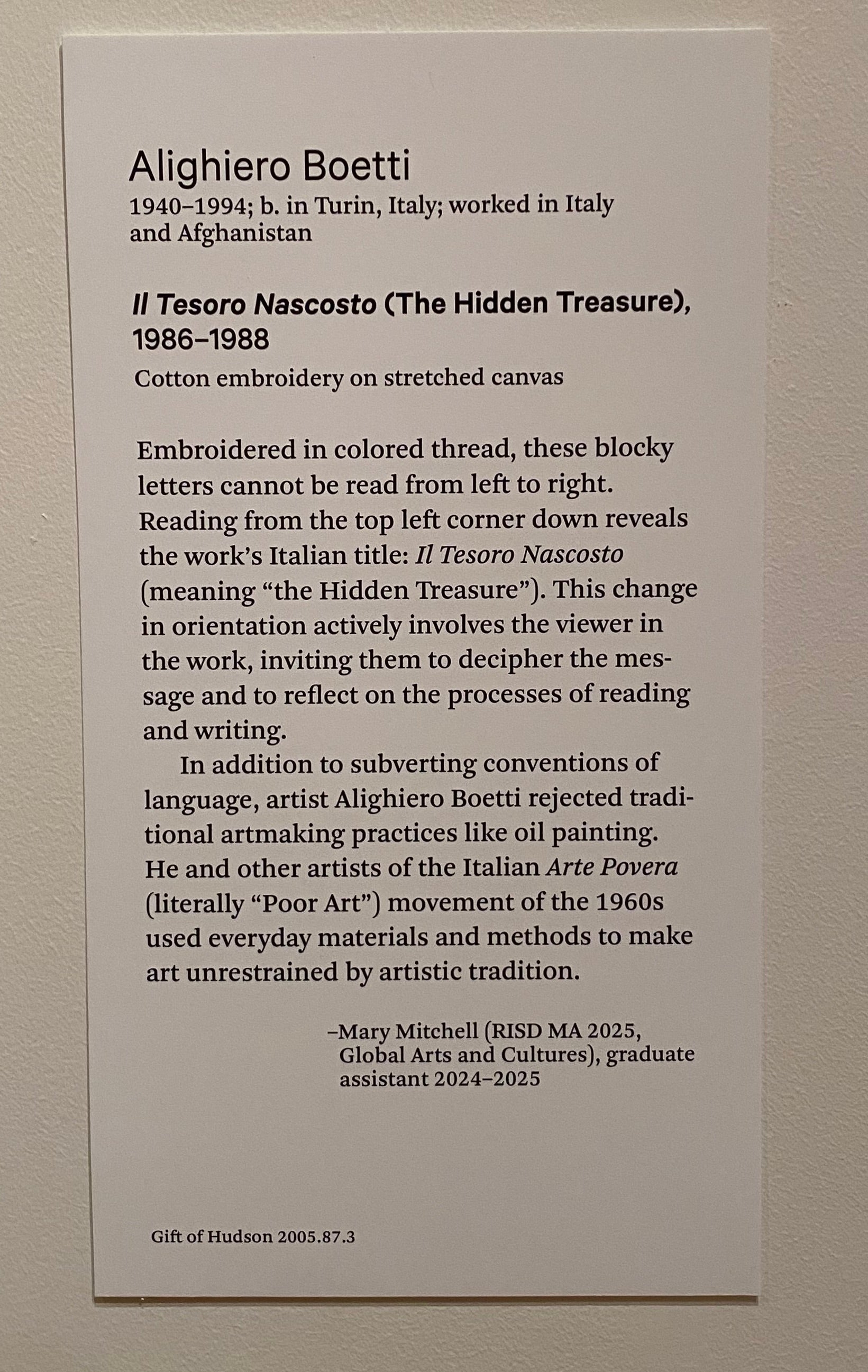 A label affixed to a white wall with two short paragraphs featuring biographical information about the artist, the title of the work, and a list of materials used. Below are two paragraphs of interpretive description that explain the viewer's involvement through reading the work's message and details about Boetti's practice. A signature credits a RISD grad student as the author. 