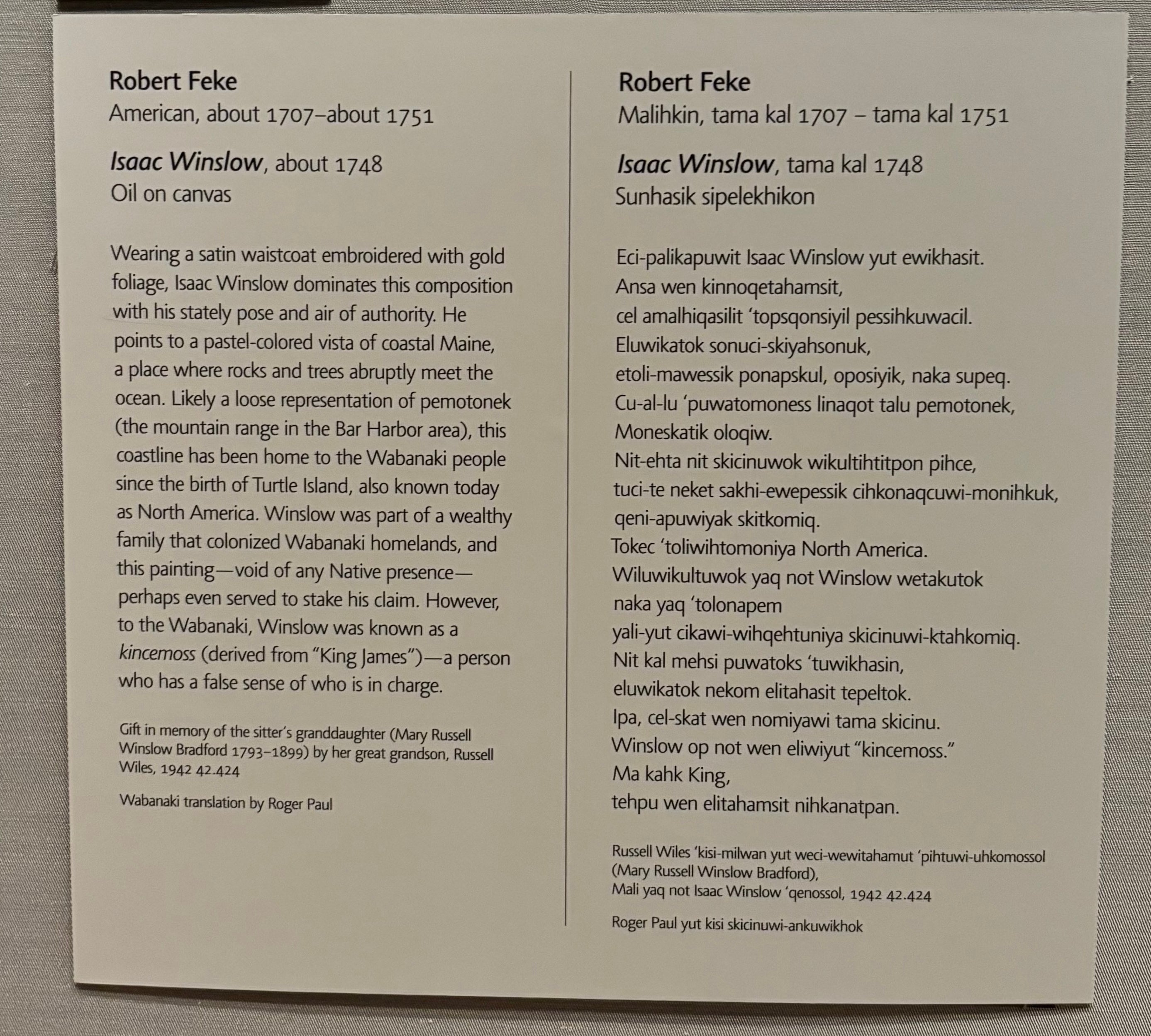 A gray label affixed to a gray wall, divided into two sections by a vertical line. On the left, biographical information about the artist, details about the painting, and a paragraph of interpretation are written in English. On the right, the same information is written in Wabanaki. 
