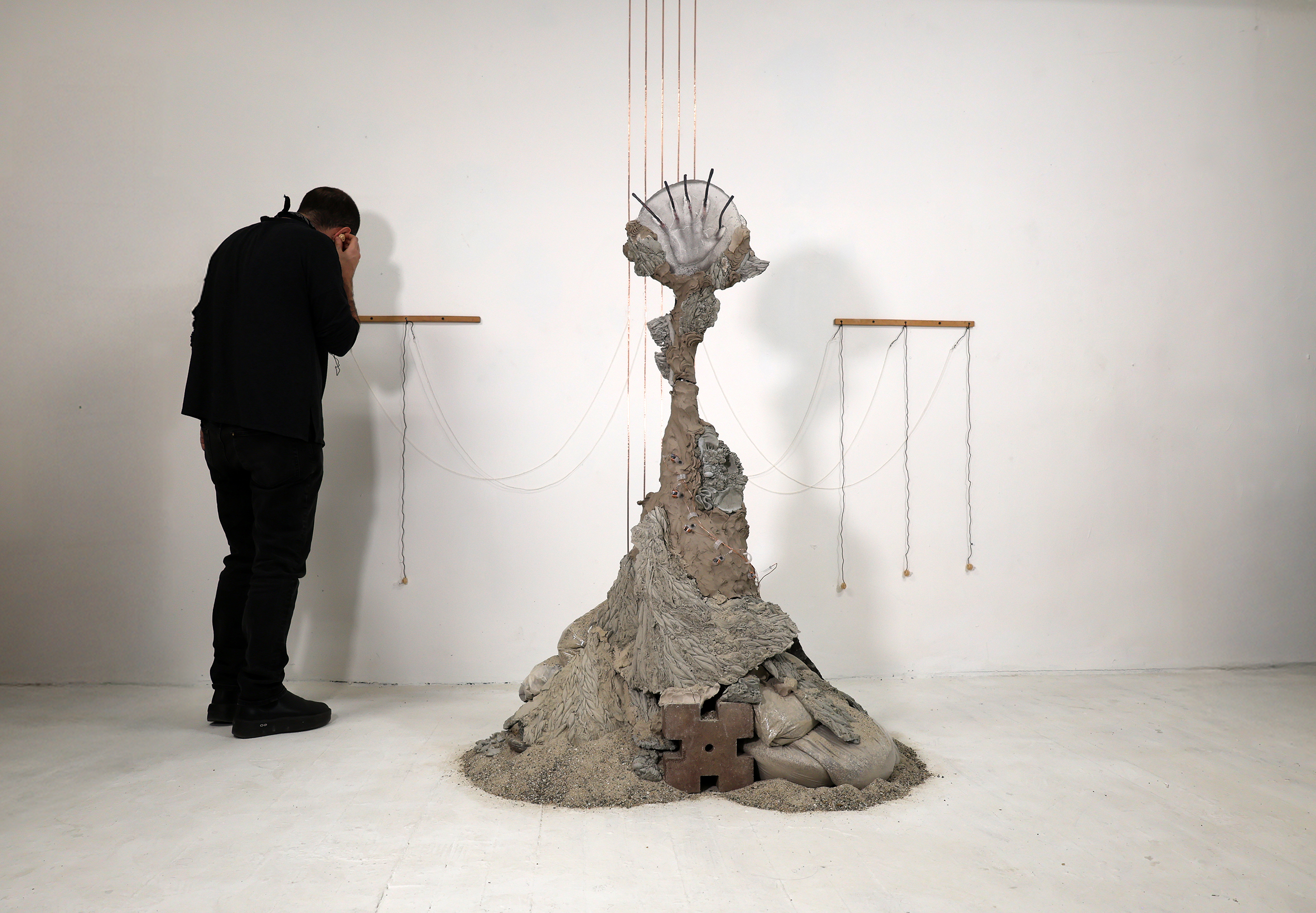 figure listening to mound of clay, concrete rubble, glass hand, copper wire around and up to the ceiling. sand on floor.