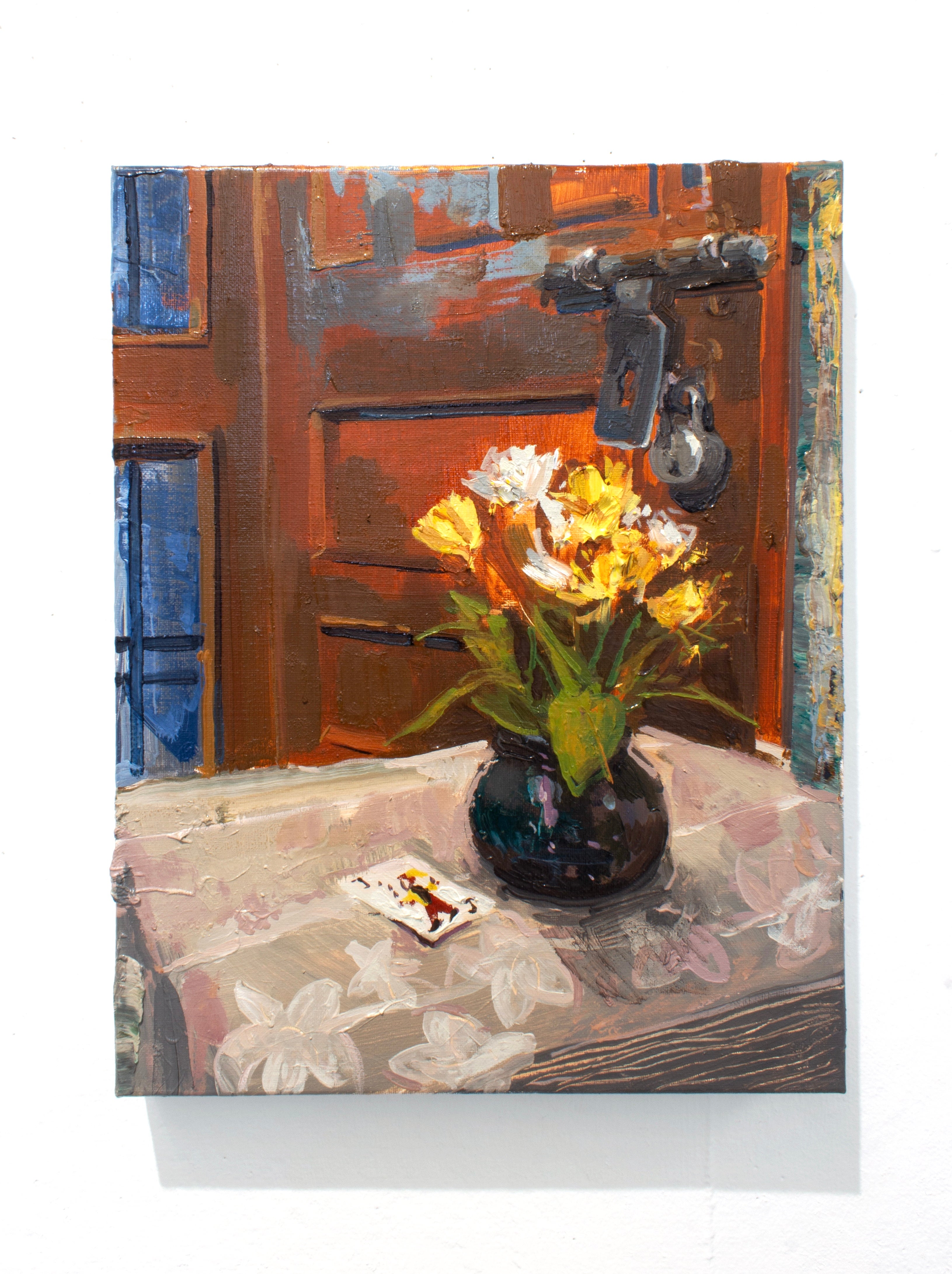 Yellow plastic flowers in a dark vase next to a joker card on a table with white floral tablecloth. Behind the vase is a wooden door with a lock.