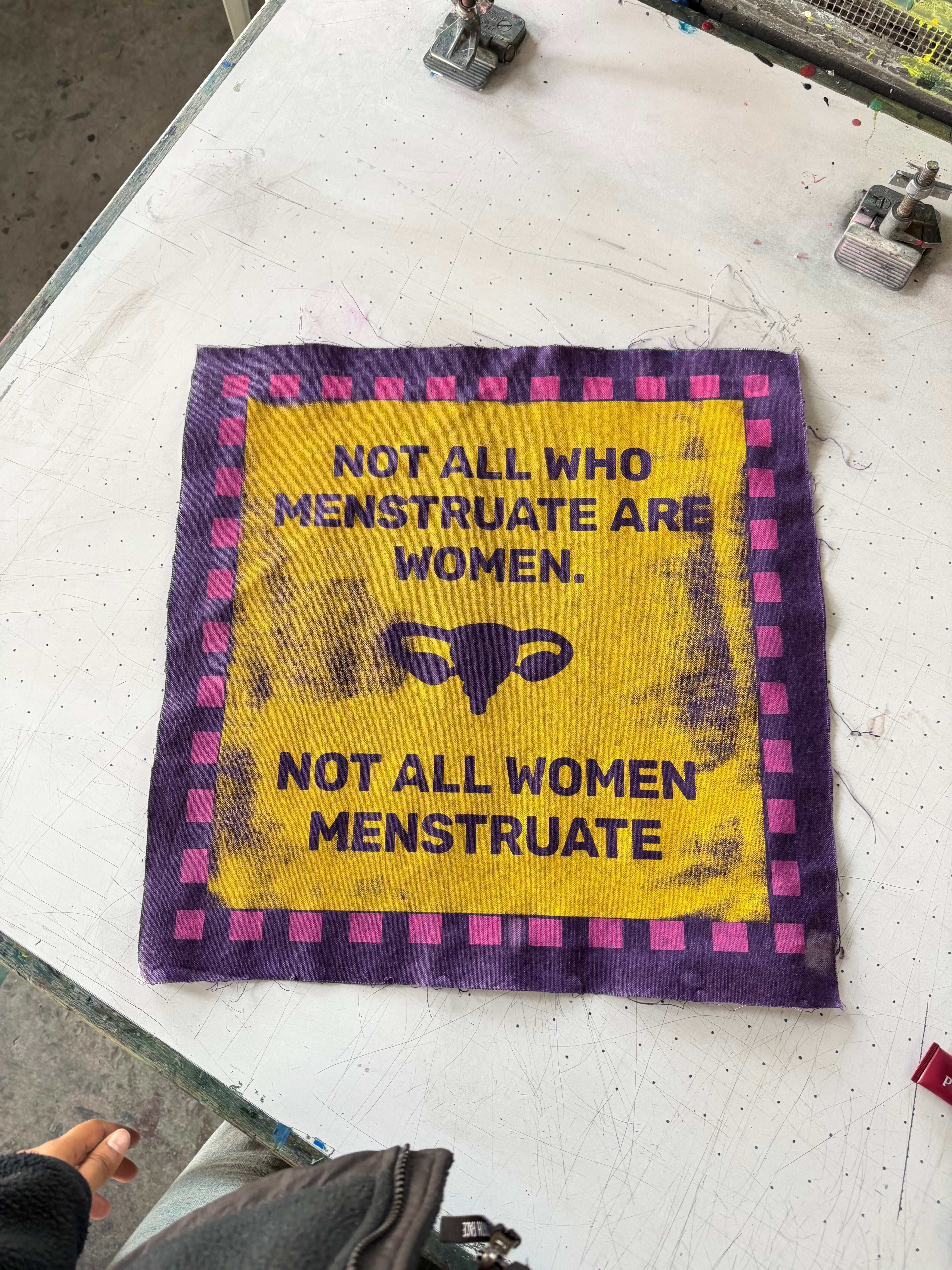 "Screen print on fabric, yellow, purple & pink A bold, graphic patch printed in purple on a golden-yellow field with a pink-and-purple checkered border. The text declares: ""Not All Who Menstruate Are Women. Not All Women Menstruate."" — flanked by a uterus icon. Photographed fresh off the print table."