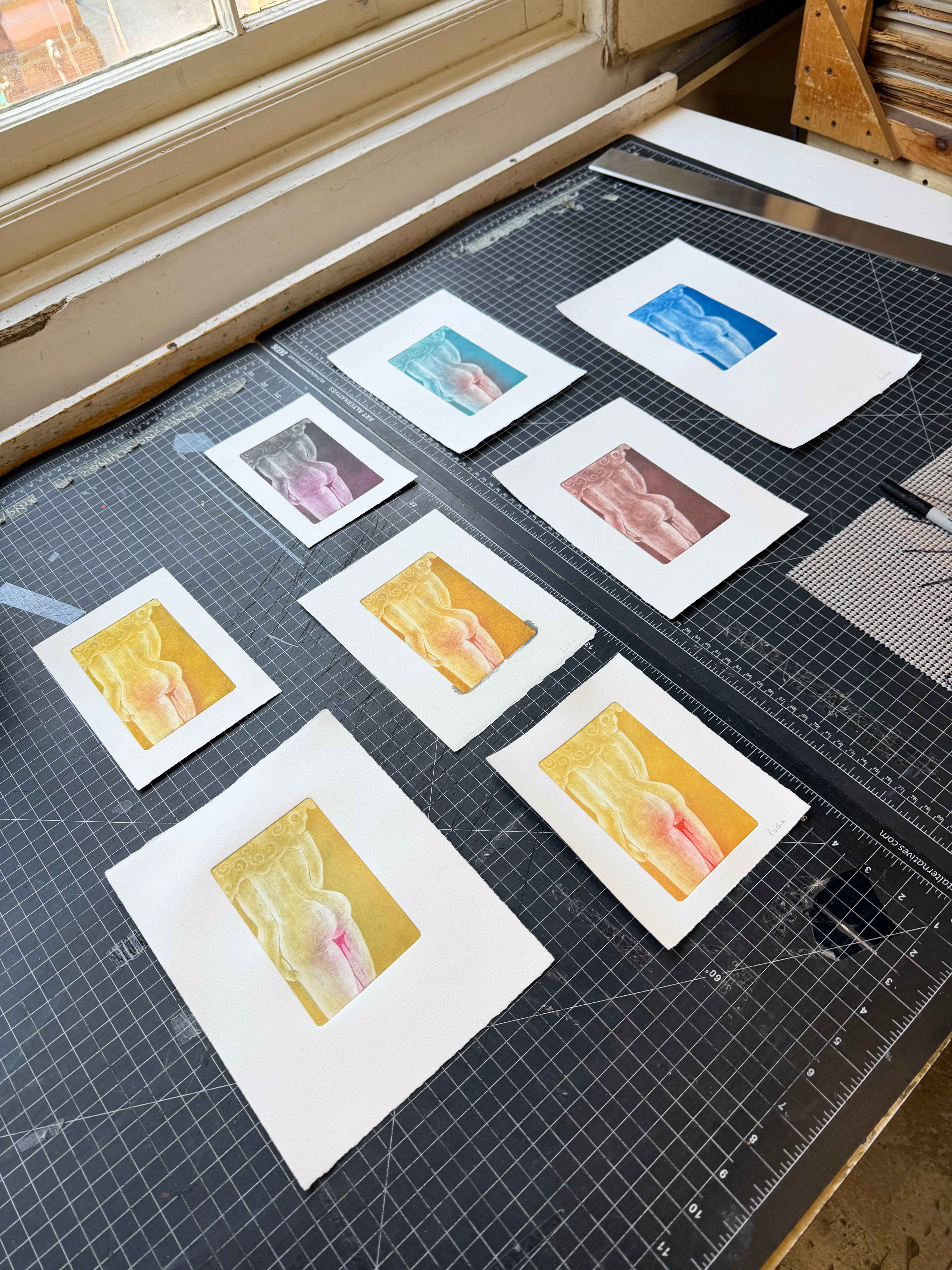 "Monoprints / relief prints on white paper, studio spread Seven prints laid out across a cutting mat, each featuring the same torso figure printed in a different colorway — teal, cobalt blue, mauve, warm brown, soft gold, and golden-rose. A studio documentation of an edition in progress, exploring how color shifts the emotional register of the same image."