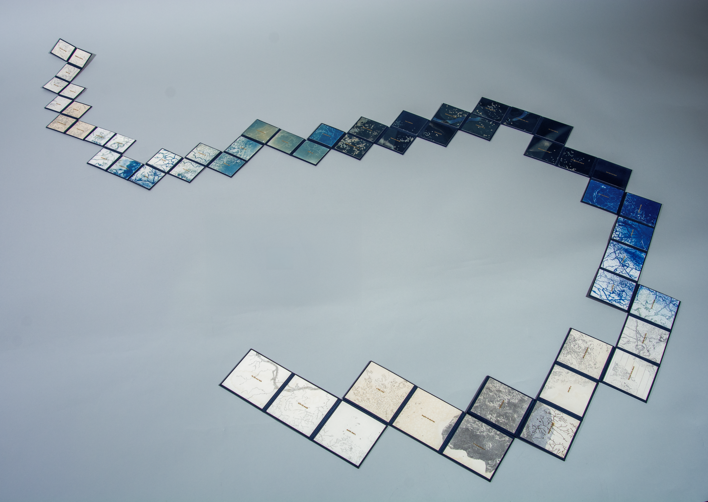 Squares of printed material ranging in color and tones of tan to dark blue are connected in a curved shape, receding in size