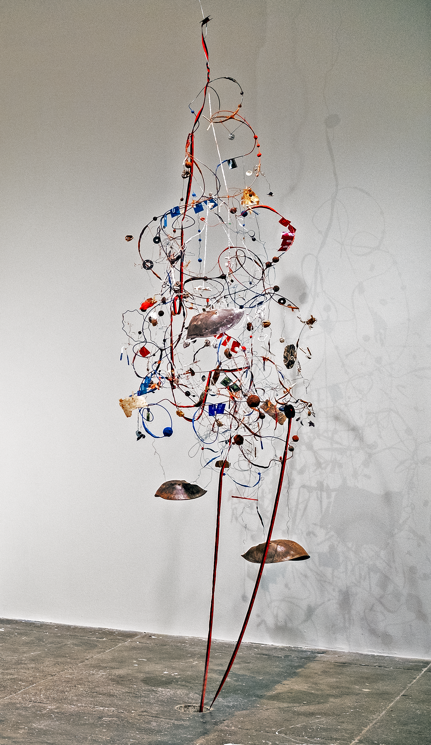 Found objects connected by wire and string, suspended from the ceiling. There is a black and red typewriter ribbon that unravels from top of the piece, disappearing through a hole in the floor.