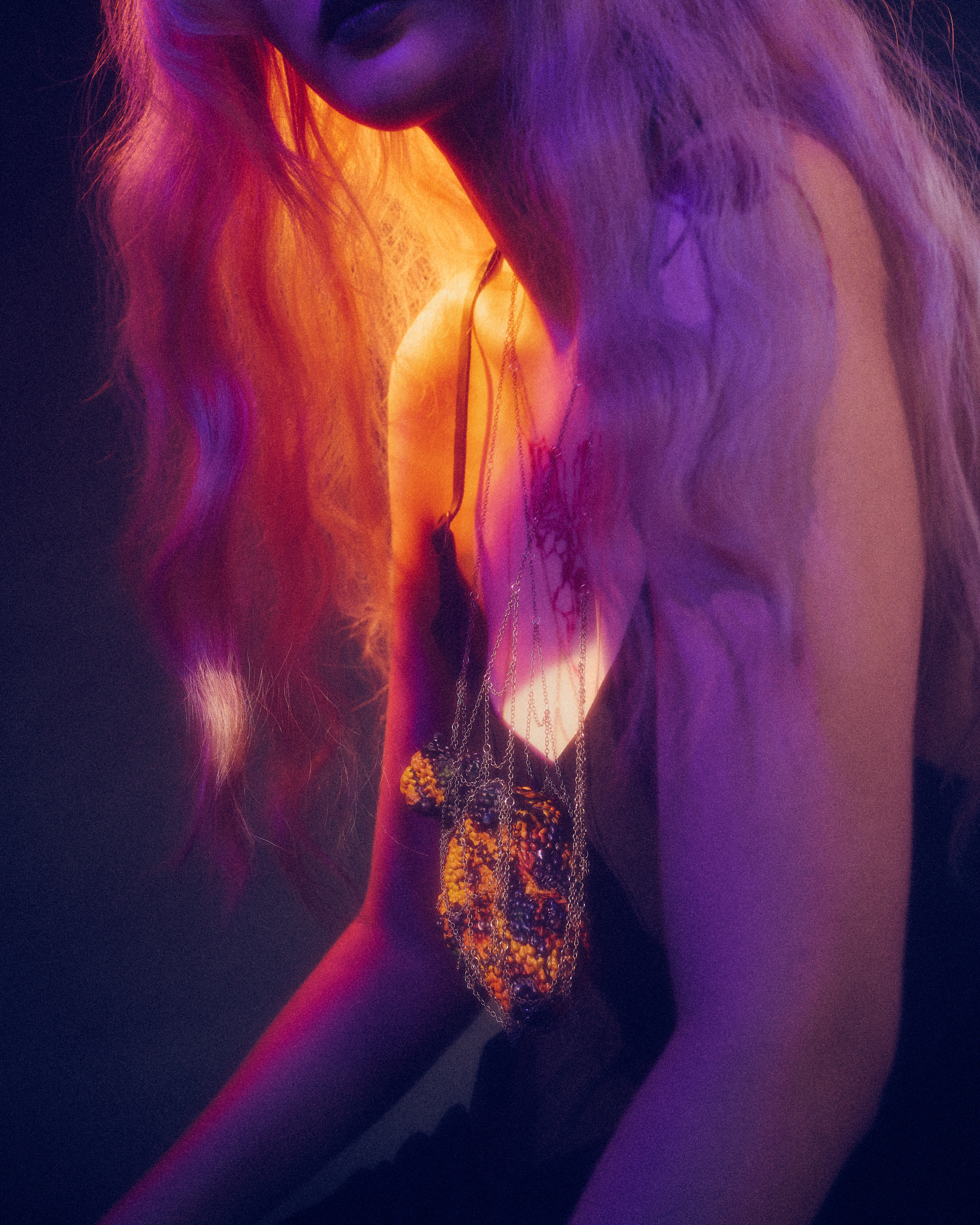 Close-up of a light-skinned figure in a black dress, angled left, wearing layered chains suspending a textured orange-brown heart-like pendant; saturated purple and warm amber lighting create an intimate, moody atmosphere.