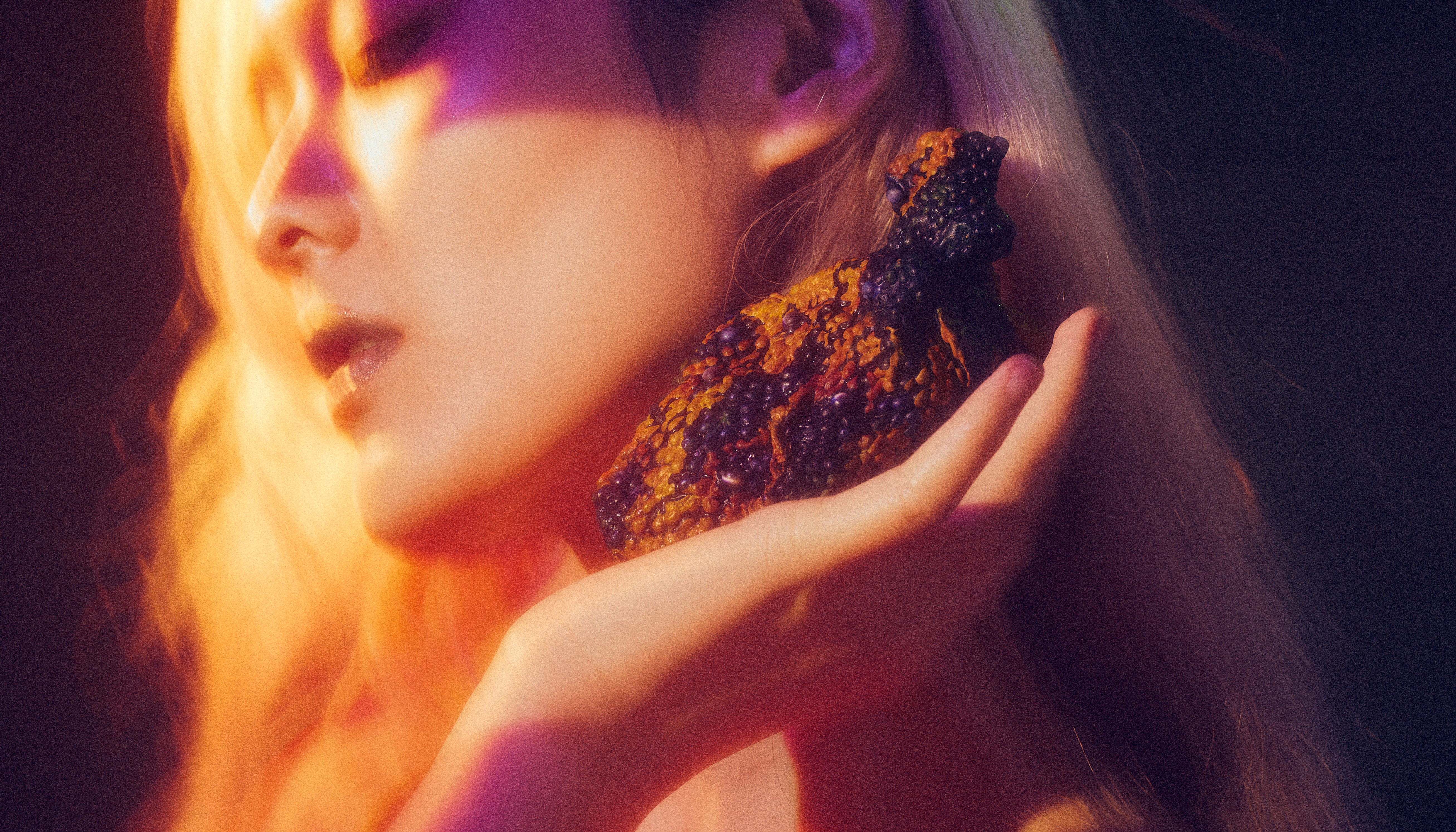Textured orange-brown heart-like object rests in a hand at right, raised to the ear; clustered dark nodules catch warm amber light, fading into deep purple shadow, creating an intimate, tactile focus.