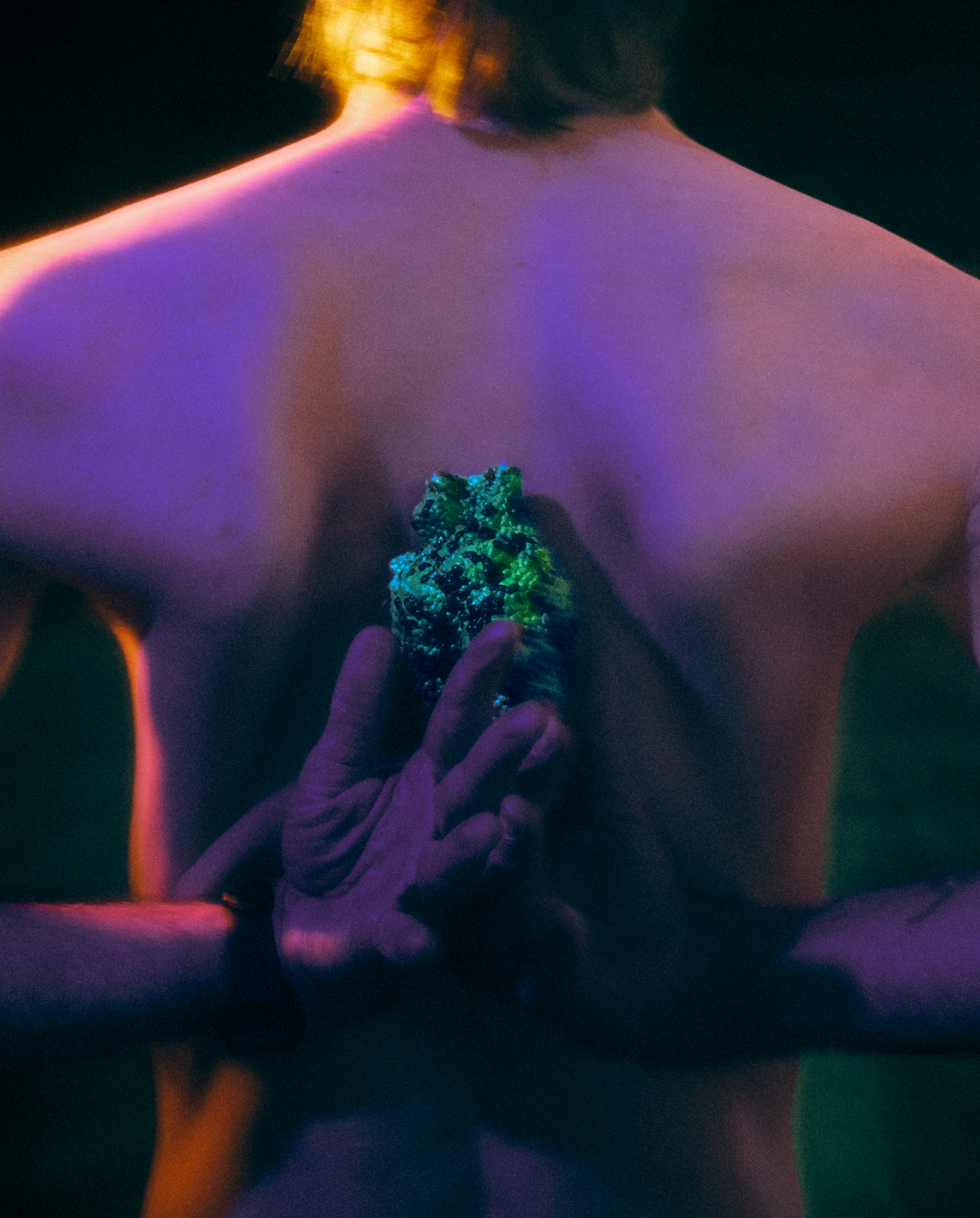 Green, textured heart-like object is pressed at center against a bare back, held by two hands from below; cool purple and deep blue shadows contrast with a warm yellow glow at the neck, creating tension.
