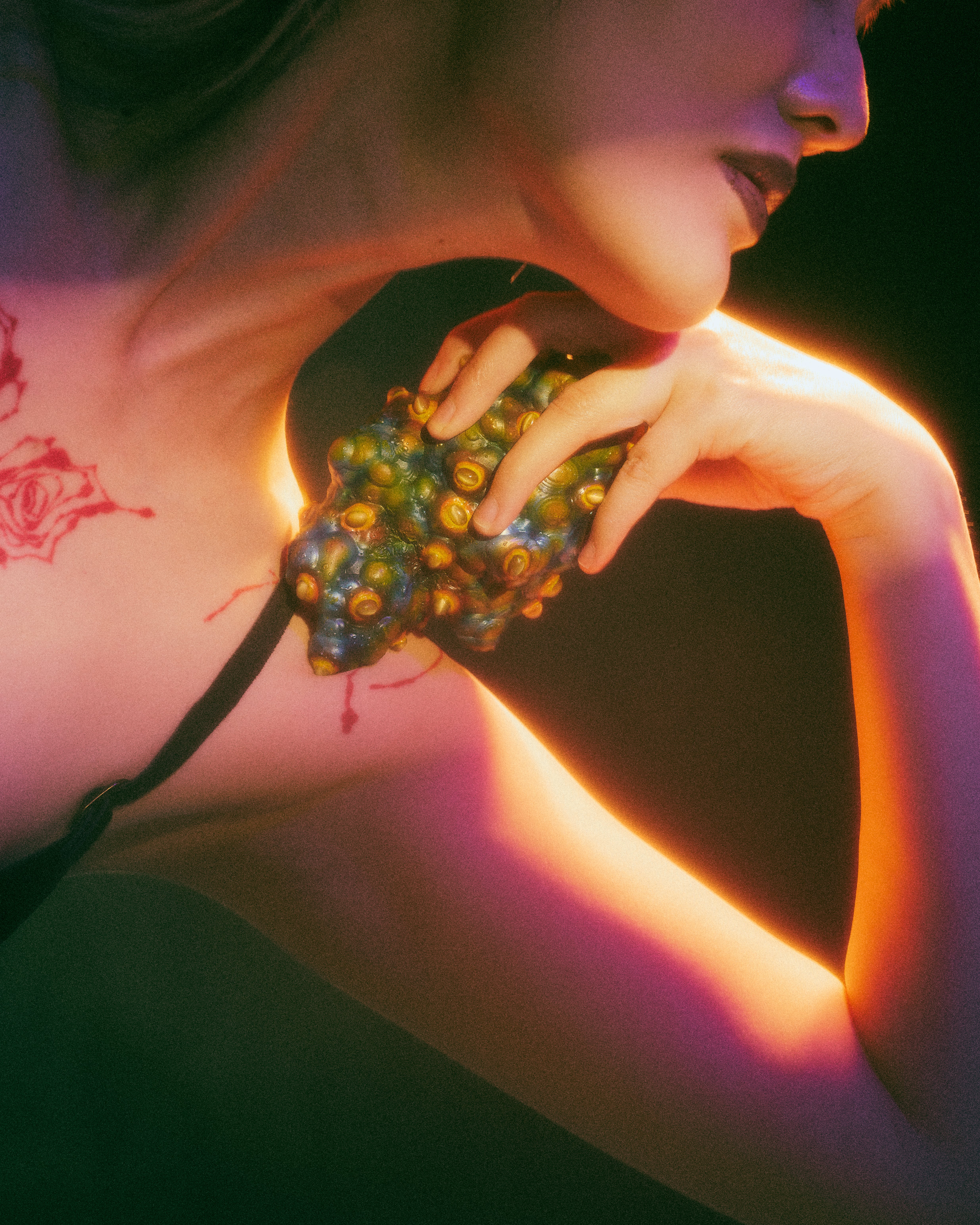 Multicolored, textured heart-like object studded with raised cat’s eye stones rests on the shoulder, held by fingers; warm amber light highlights its surface while deep purple and green shadows create an intimate, tense mood.