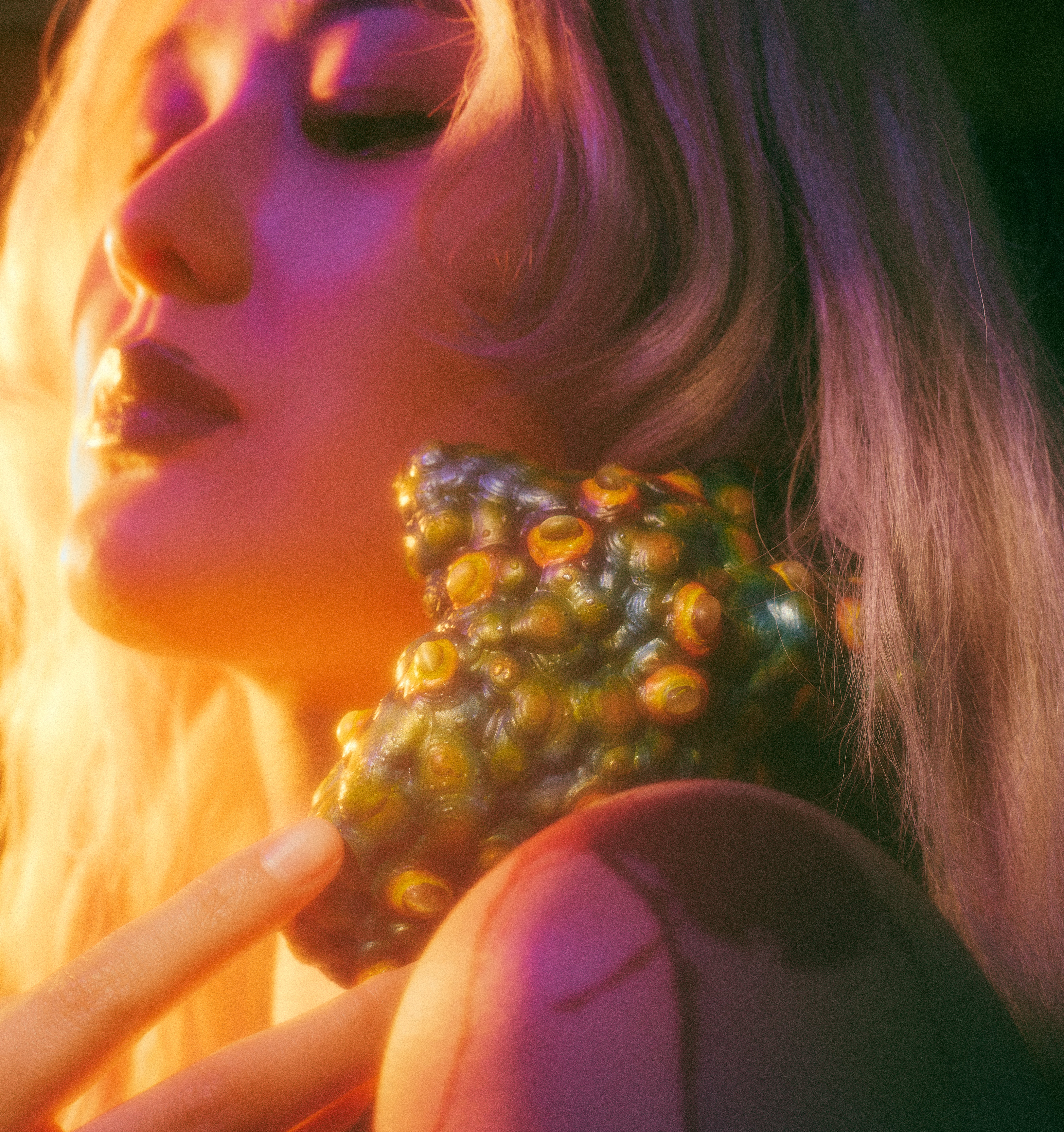 Multicolored, textured heart-like object studded with raised cat’s eye stones rests at the neck and shoulder, touched by fingers; warm amber light highlights its surface while deep purple-green shadows create an intimate, tense mood.