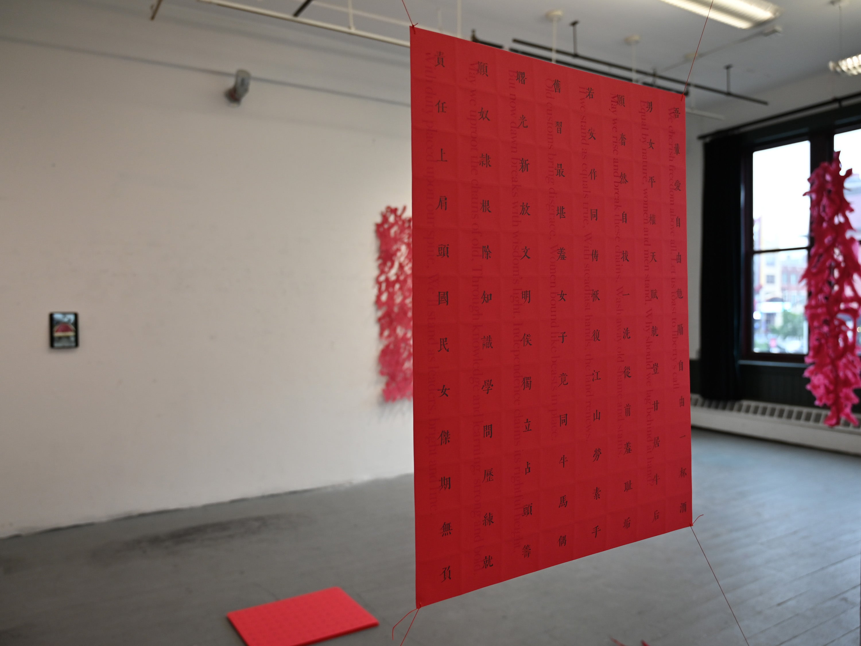 Red hanging paper sheet with vertical text, suspended in a gallery with red paper installations in the background.