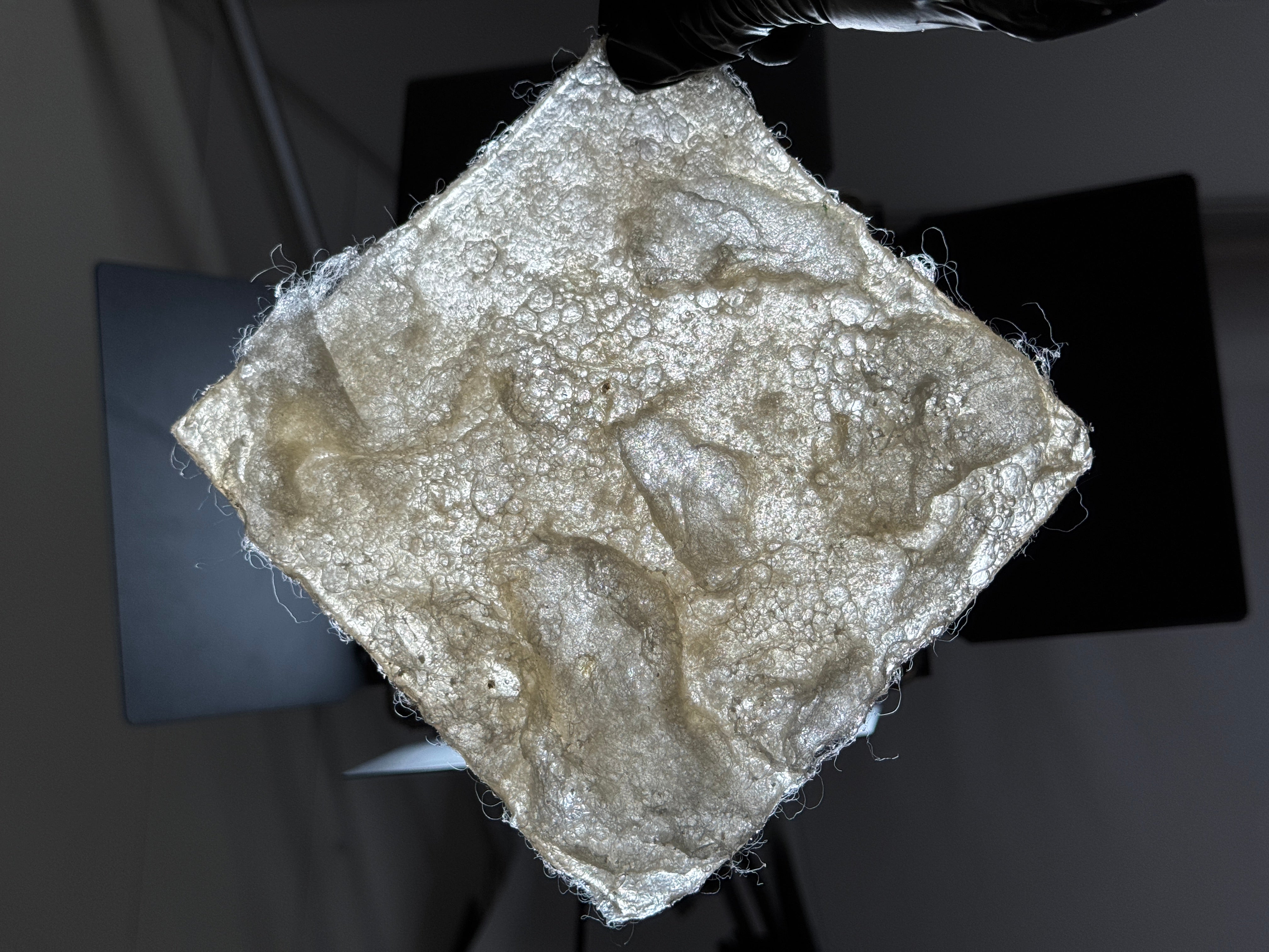 A bioplastic made primarily from wool and gelatin. 6 inches by 6 inches. Under this recipe and processing method, the surface develops large bubbles that can be pressed and spring back, forming a cross-linked network structure internally. It is translucent with a slight yellowish tint.