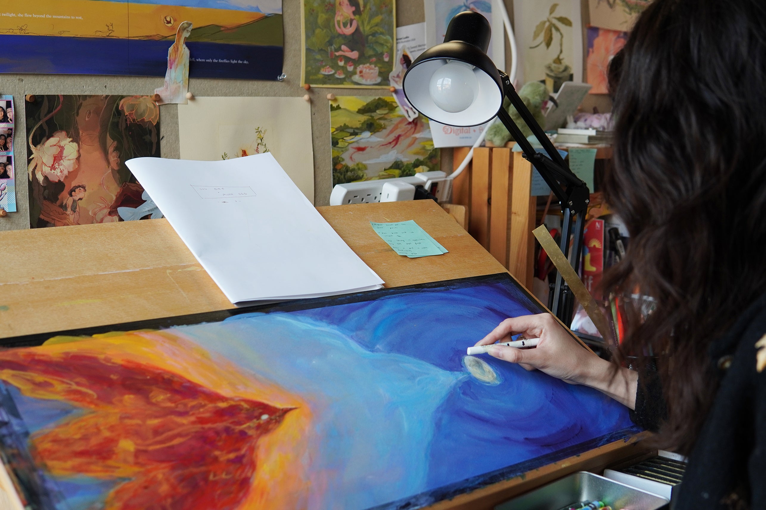 Photograph of the artist in her studio working on a picture book spread depicting a red bird flying from the left page toward a white egg against a dark blue night sky.