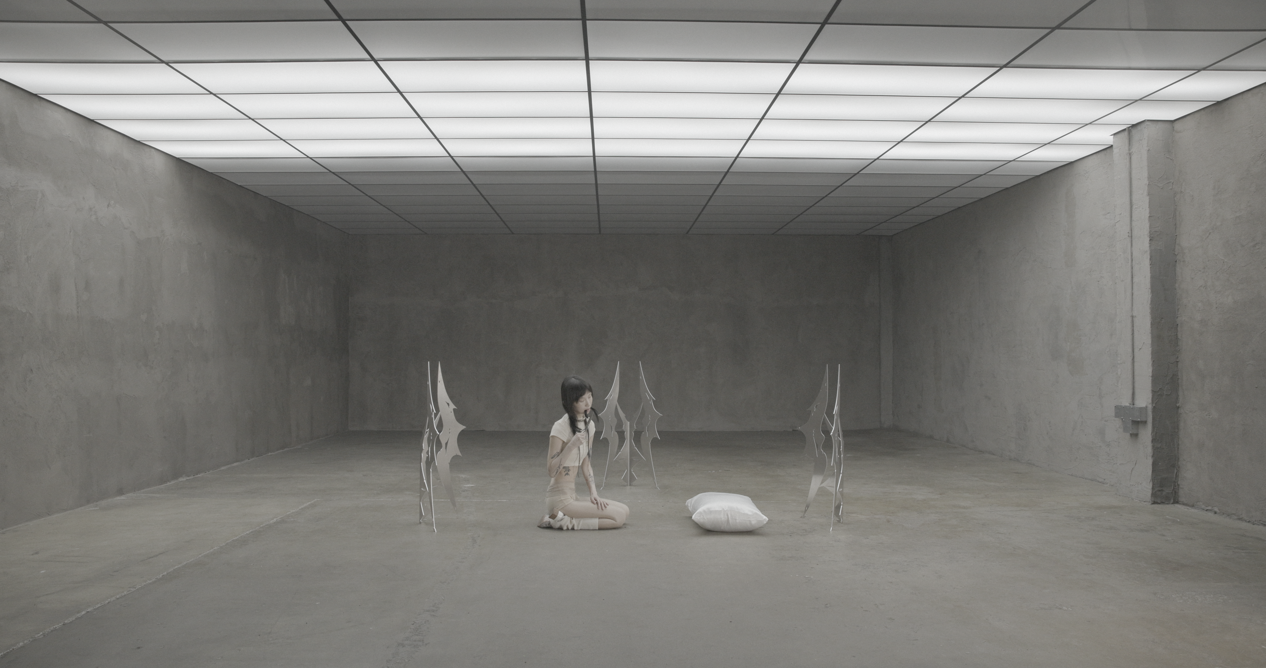 Wide shot of a person kneeling on concrete floor in a large gray room, surrounded by upright metal sculptures, holding hair near mouth; white pillow placed nearby under grid ceiling lights.