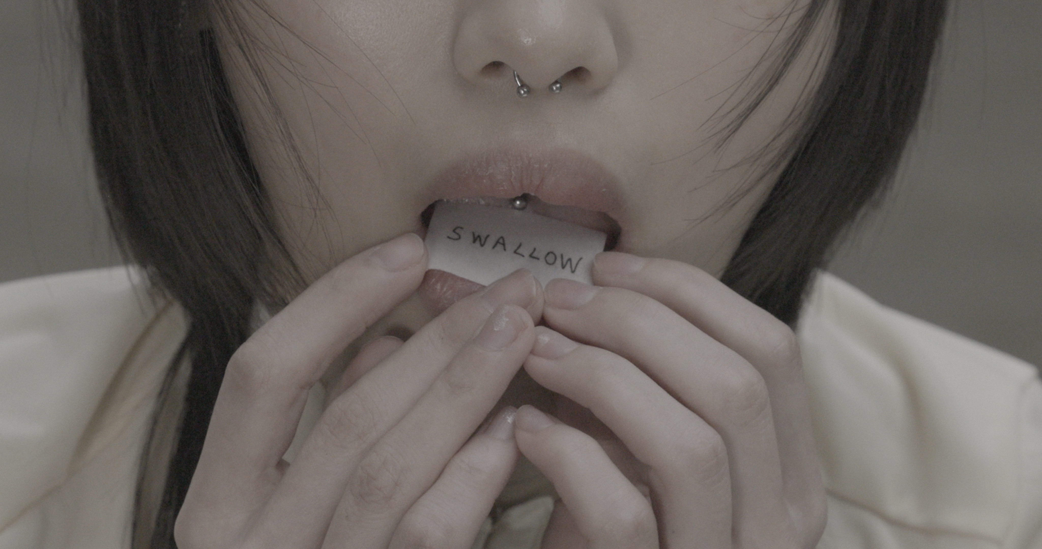 Close-up of mouth pressing a small paper labeled “SWALLOW” between lips; fingers hold edges of paper, skin and texture sharply visible.