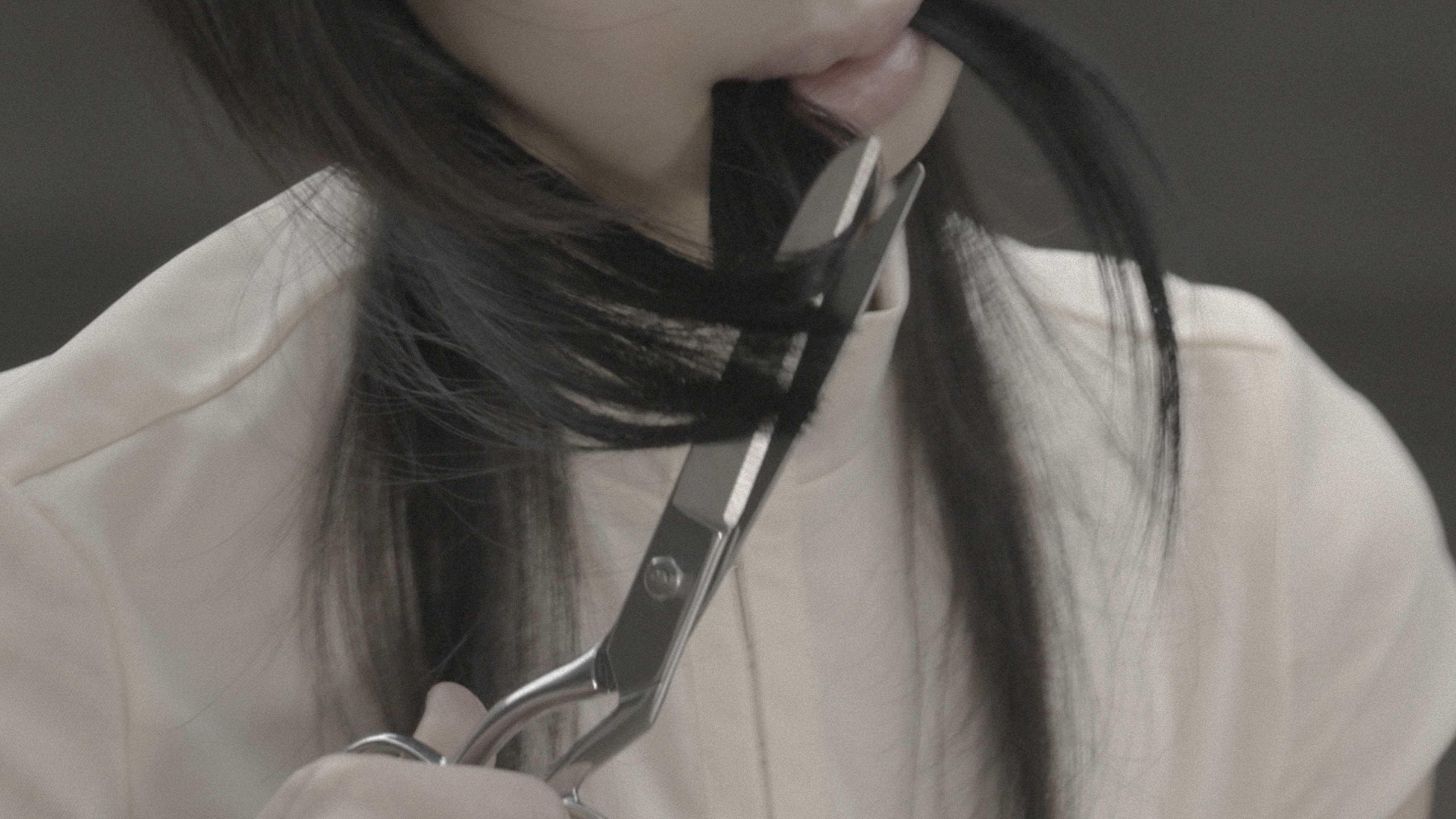 Close-up of scissors cutting a section of dark hair near the neck; strands are held taut between blades, with soft fabric clothing visible behind.
