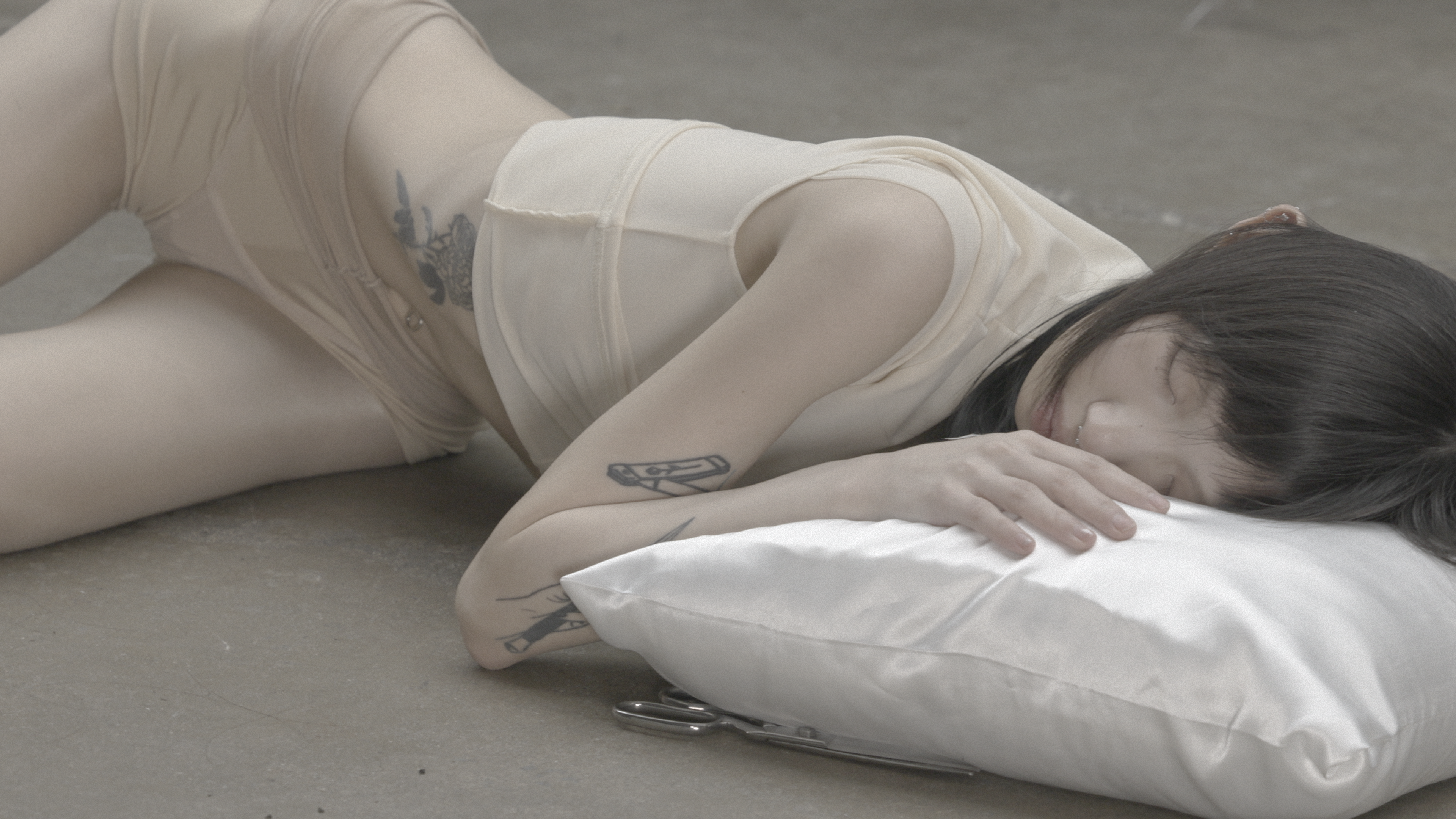 Person lying on concrete floor with head resting on a white pillow, eyes closed, body in nude-toned clothing; scissors placed beneath pillow edge, soft lighting throughout.