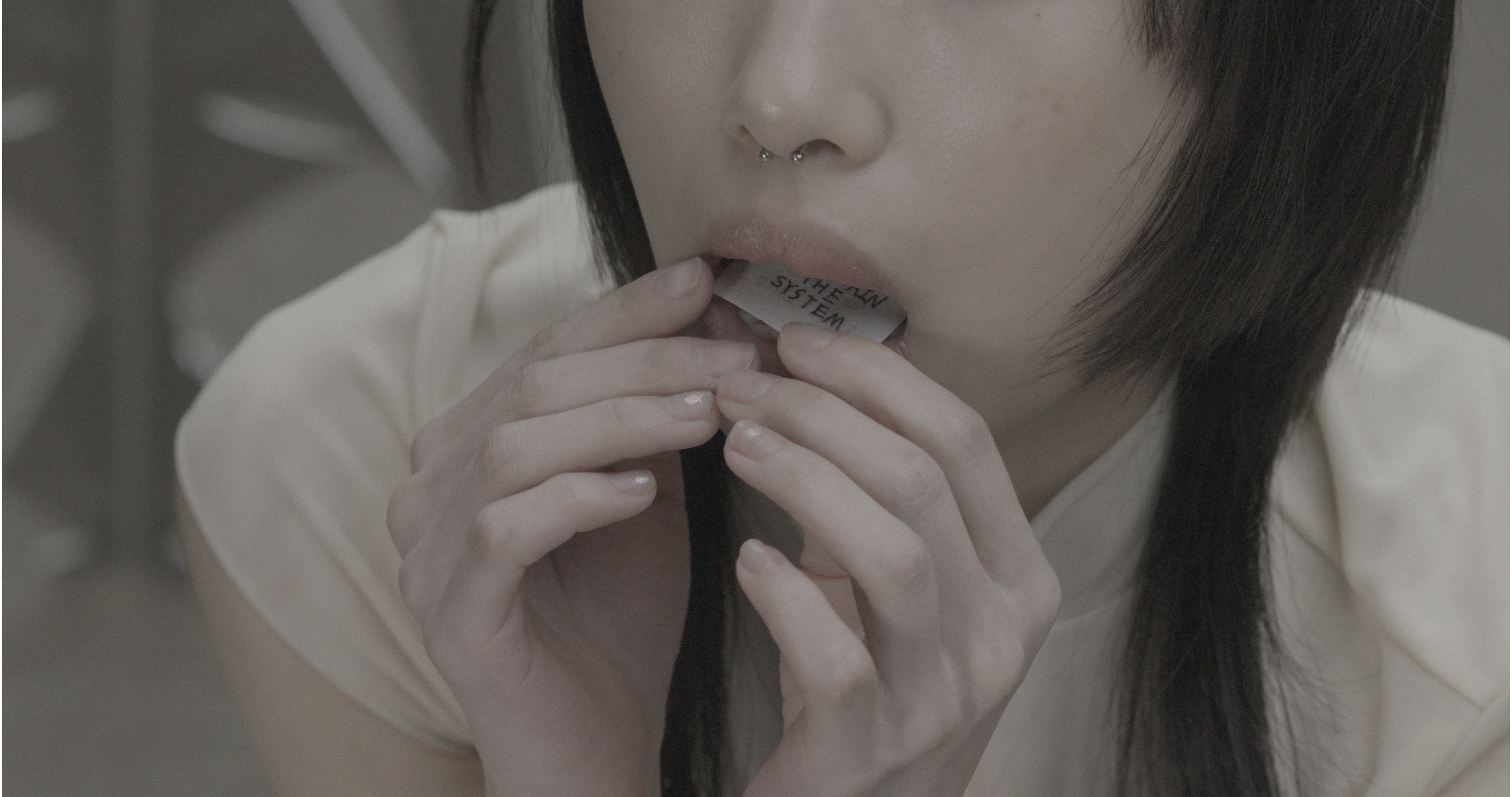 Close-up of mouth holding a small paper labeled “MAINTAIN THE SYSTEM,” fingers pressing it inward; neutral tones, hair framing face, expression controlled.