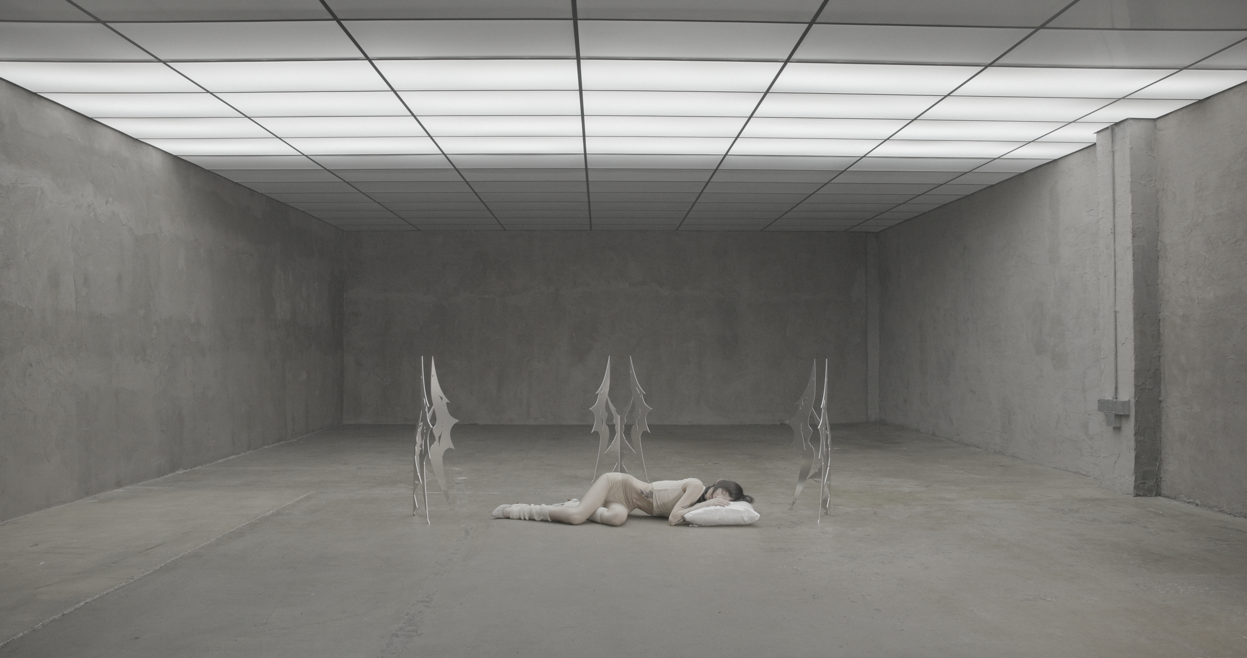 Wide shot of person lying on floor within large empty gray room, surrounded by metal sculptures, head resting on white pillow under grid ceiling lights.