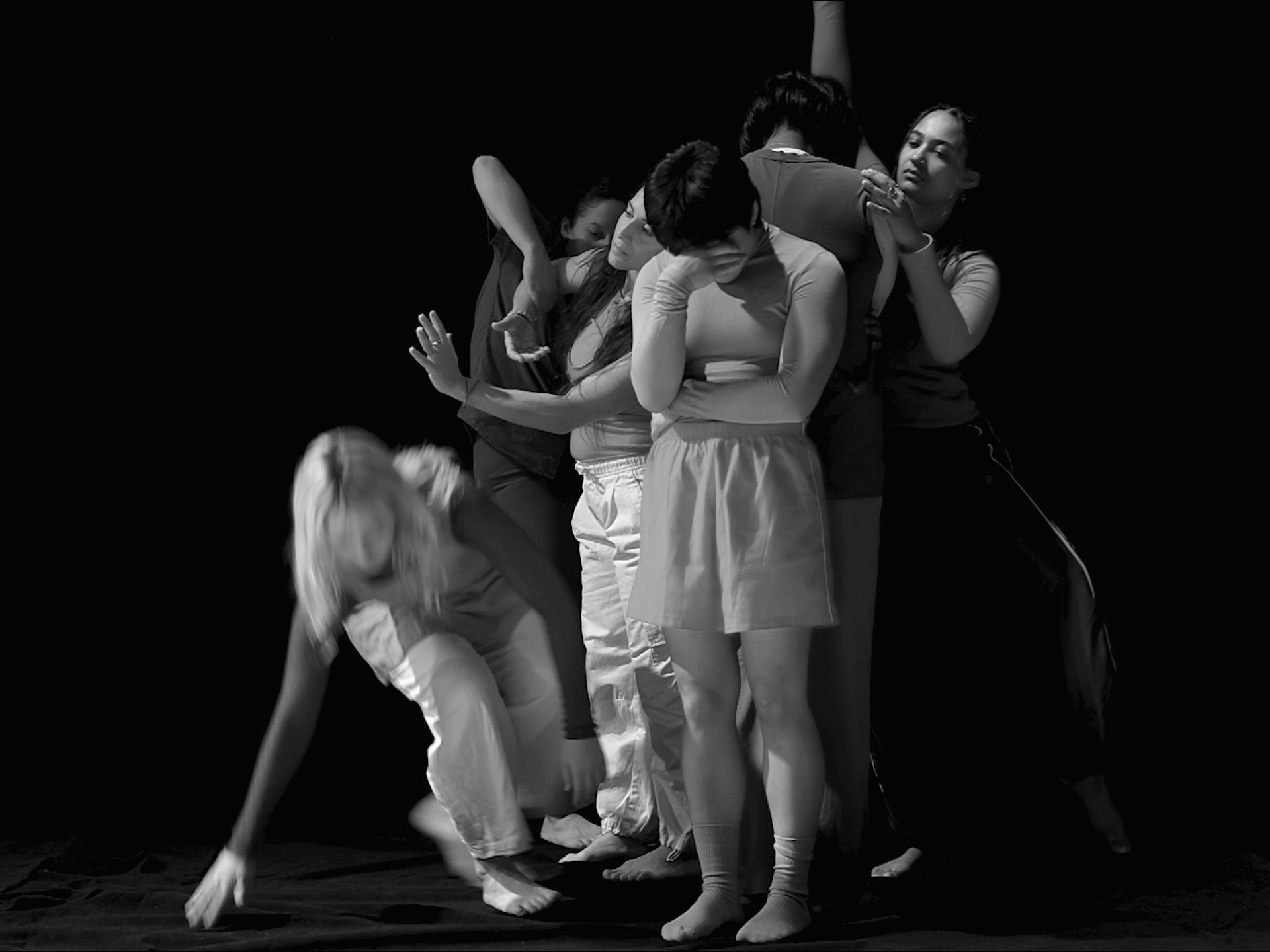 Six female and non-binary identifying dancers create various shapes with their bodies against a black background in this black and white video still.