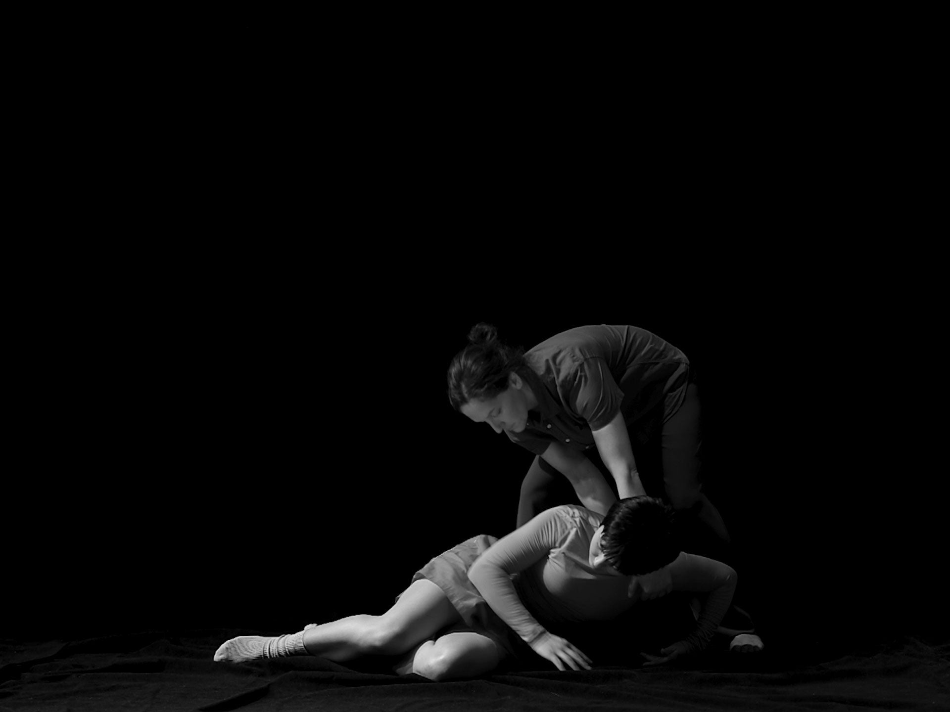 Two (female/non-binary identifying) figures are pictured against a black background in this black and white video still. 