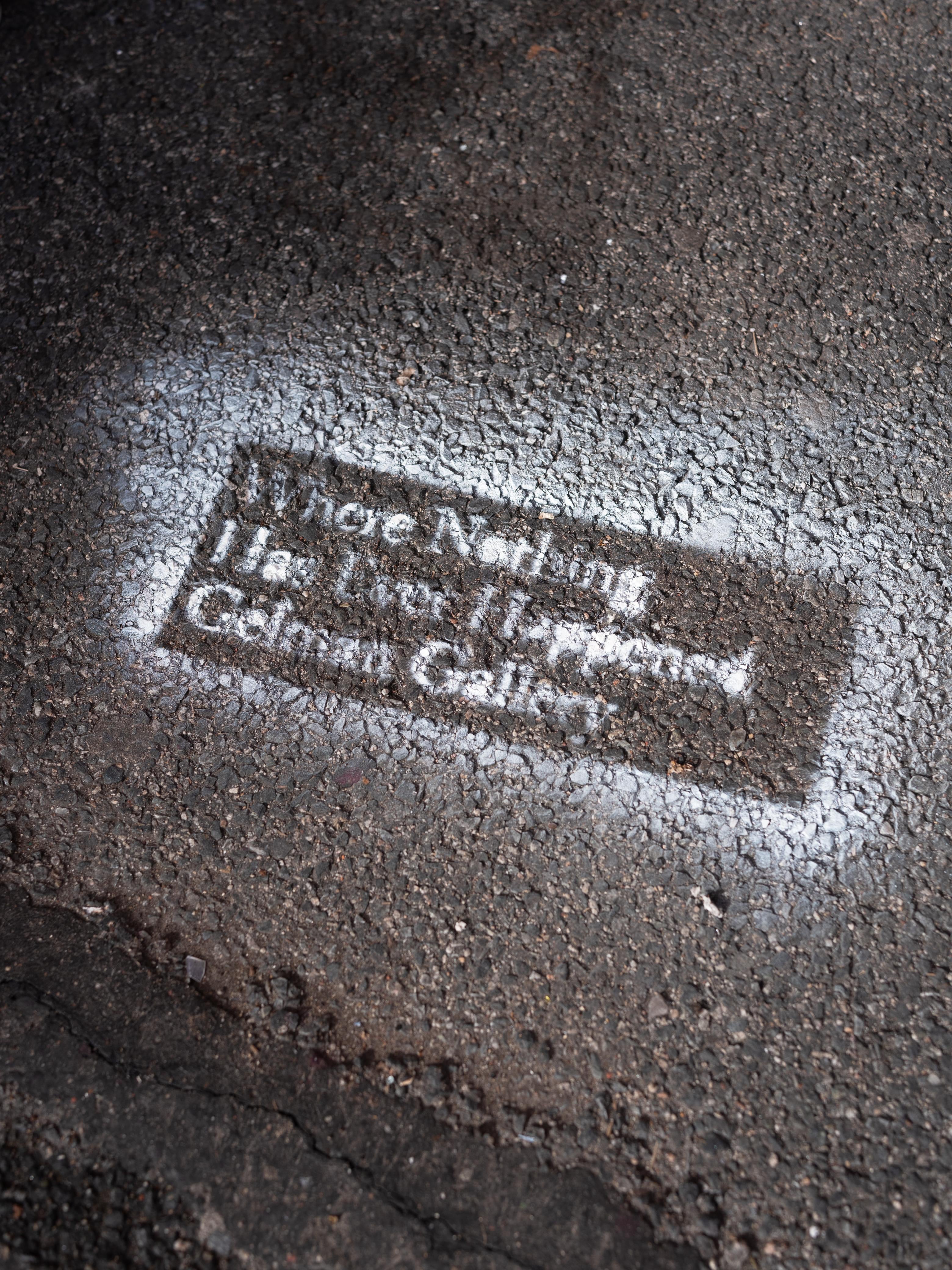 Close-up of a white stenciled text on dark, wet asphalt reading "Where Nothing Has Ever Happened / Gelman Gallery," photographed at an angle to emphasize the rough texture of the pavement.