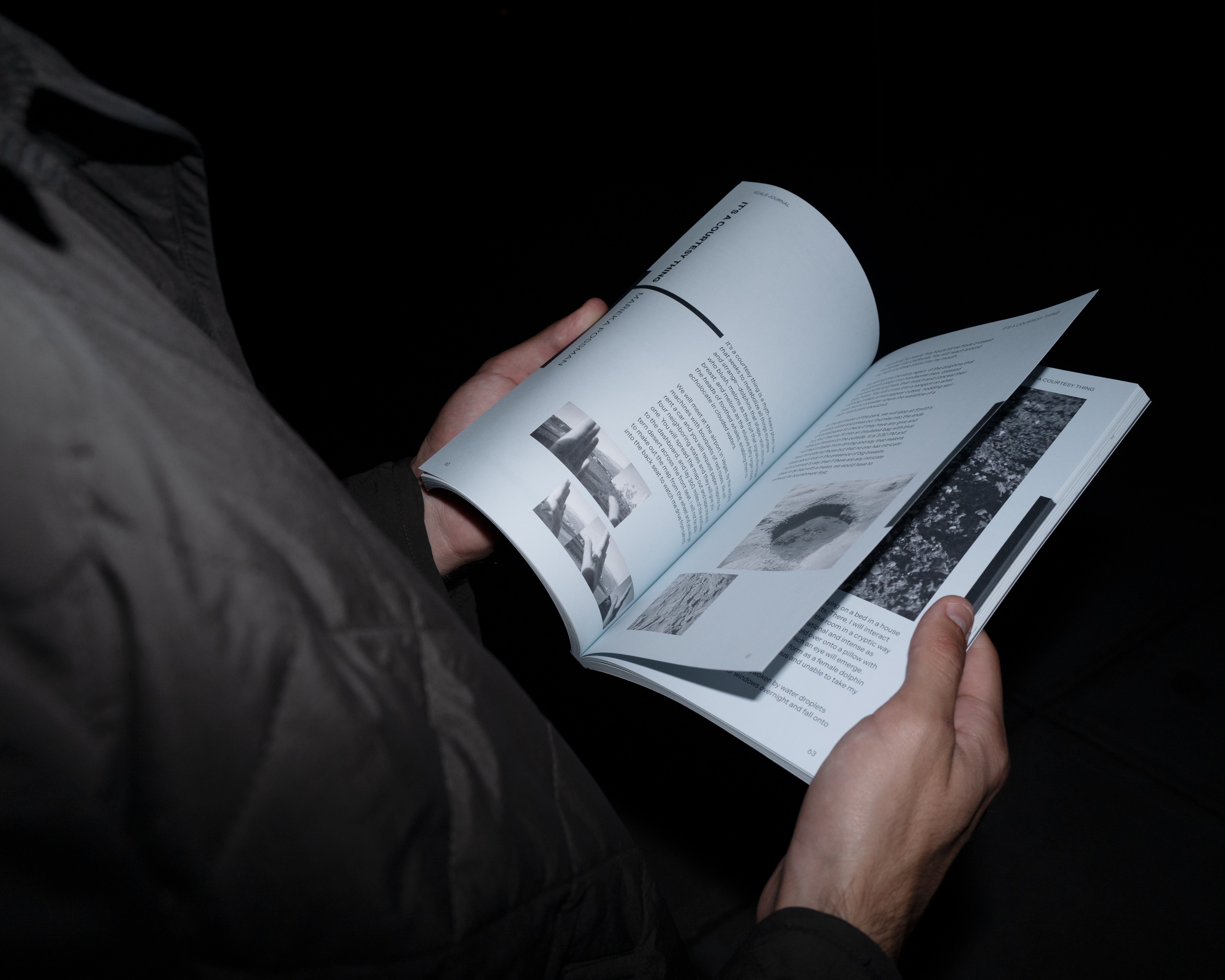 A person's hands holding an open copy of Scale Journal in a dark setting. The visible spread shows black-and-white photographs alongside body text under the section heading "It's a Courtesy Thing."