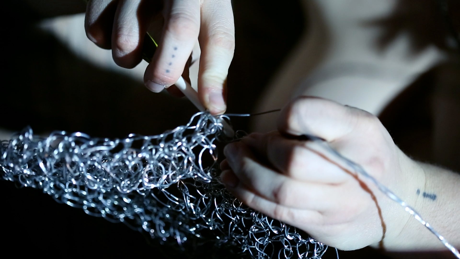 Close-up of hands crocheting thin silver wire into a tangled mesh, with one holding the hook and the other threading wire. Dramatic and focused lighting highlights metallic texture.