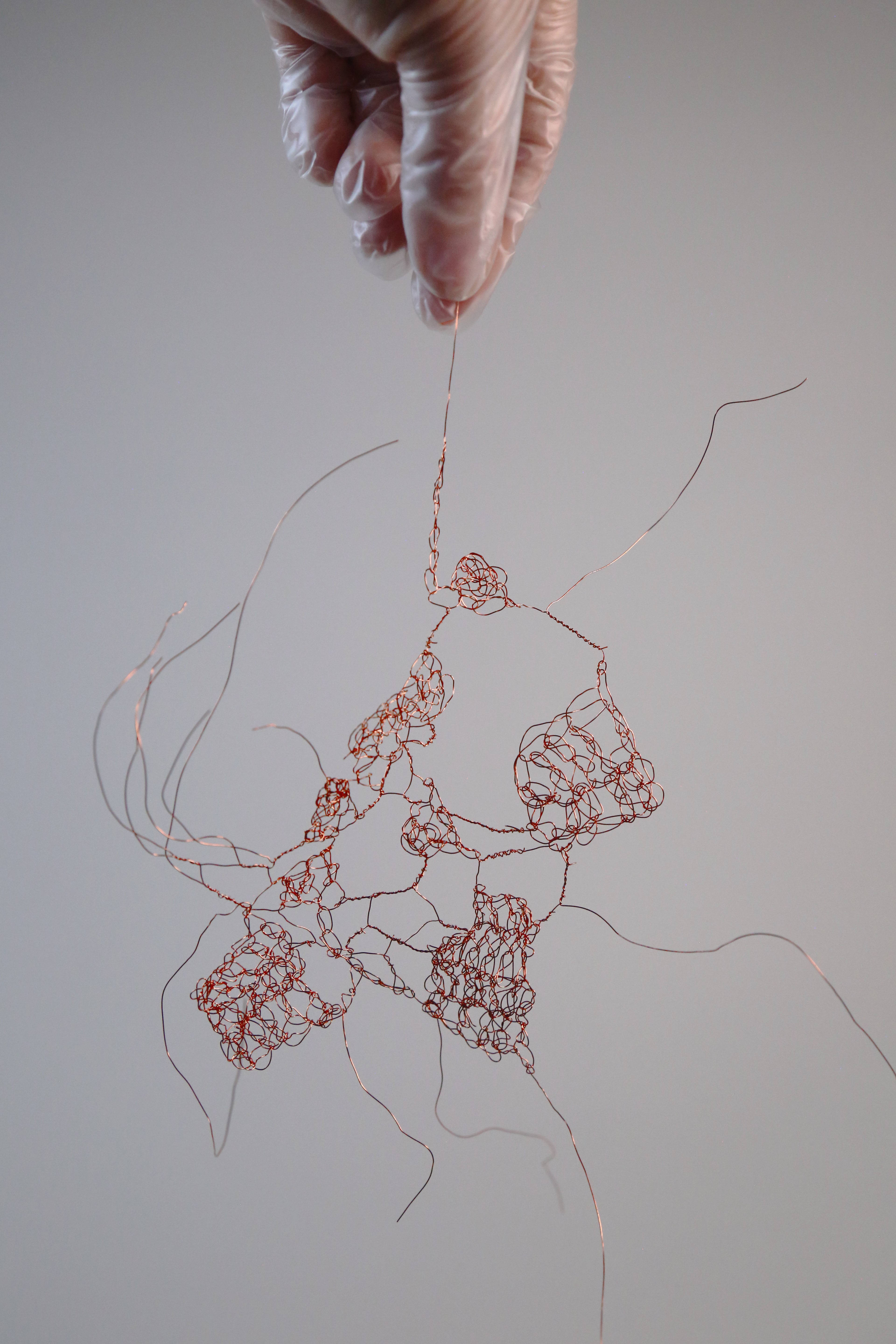 Gloved hand holds delicate copper wire sculpture, forming tangled, organic shapes suspended against a white background.