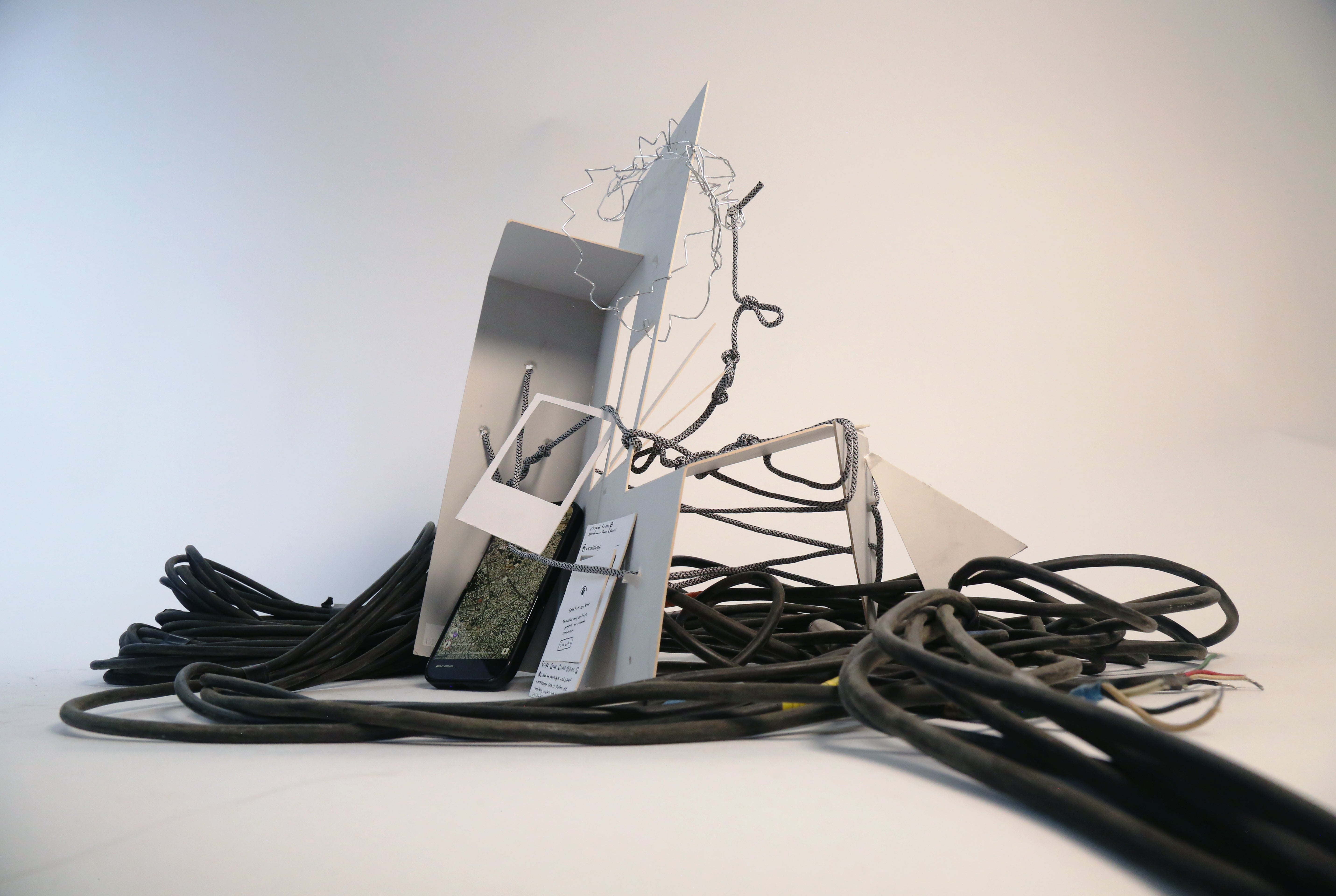 Concept model consisting of white panels, wire, and rope entangled with thick black cables, with an iphone displaying a map of Gaza, arranged against a white background.