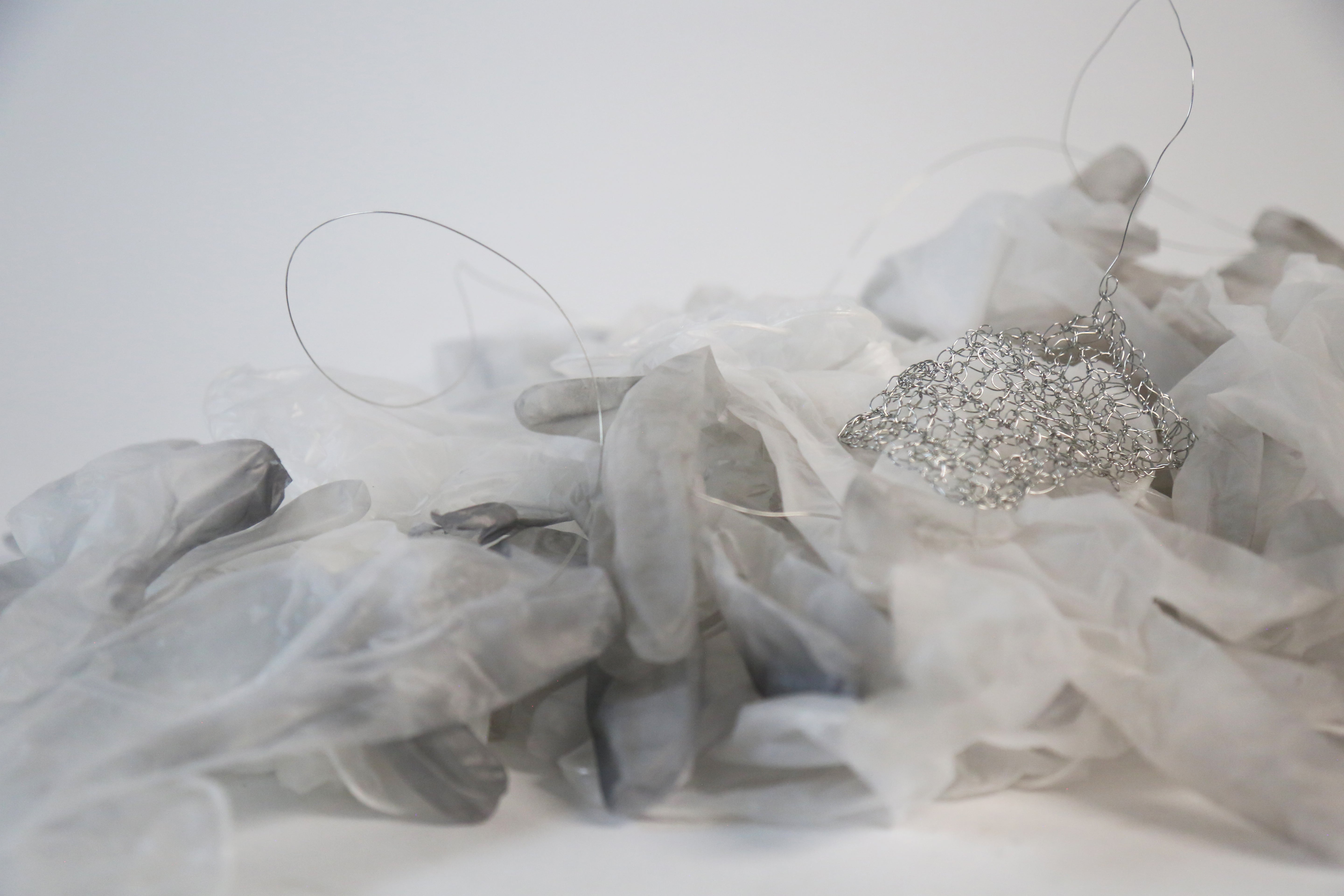 A pile of semi opaque used vinyl gloves with a delicate crocheted piece made from thin silver wire placed amidst the pile.
