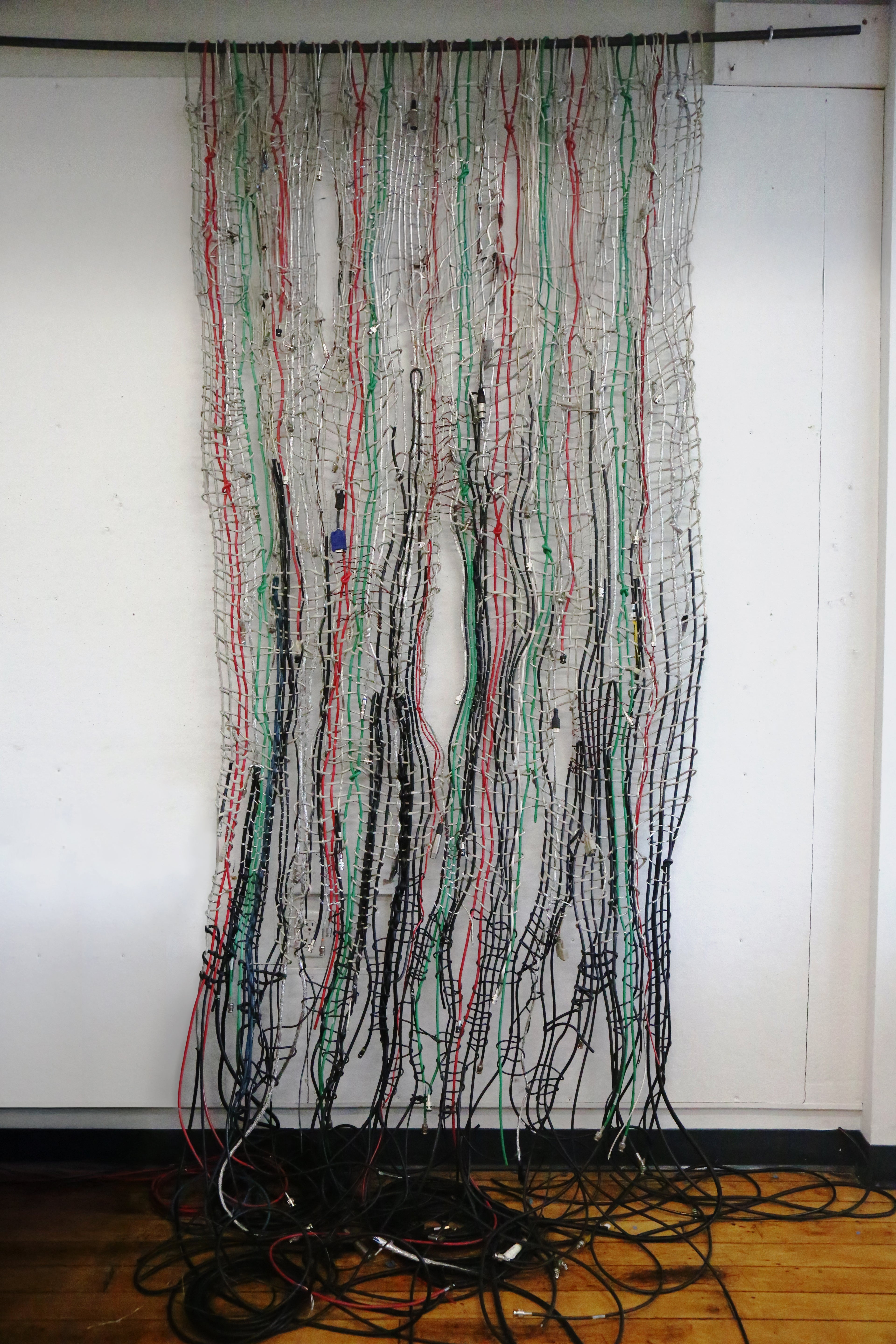 Large, dense textile of multicolored cables hanging vertically, woven with stripped metal wires against a light background, creating a chaotic, net-like pattern.