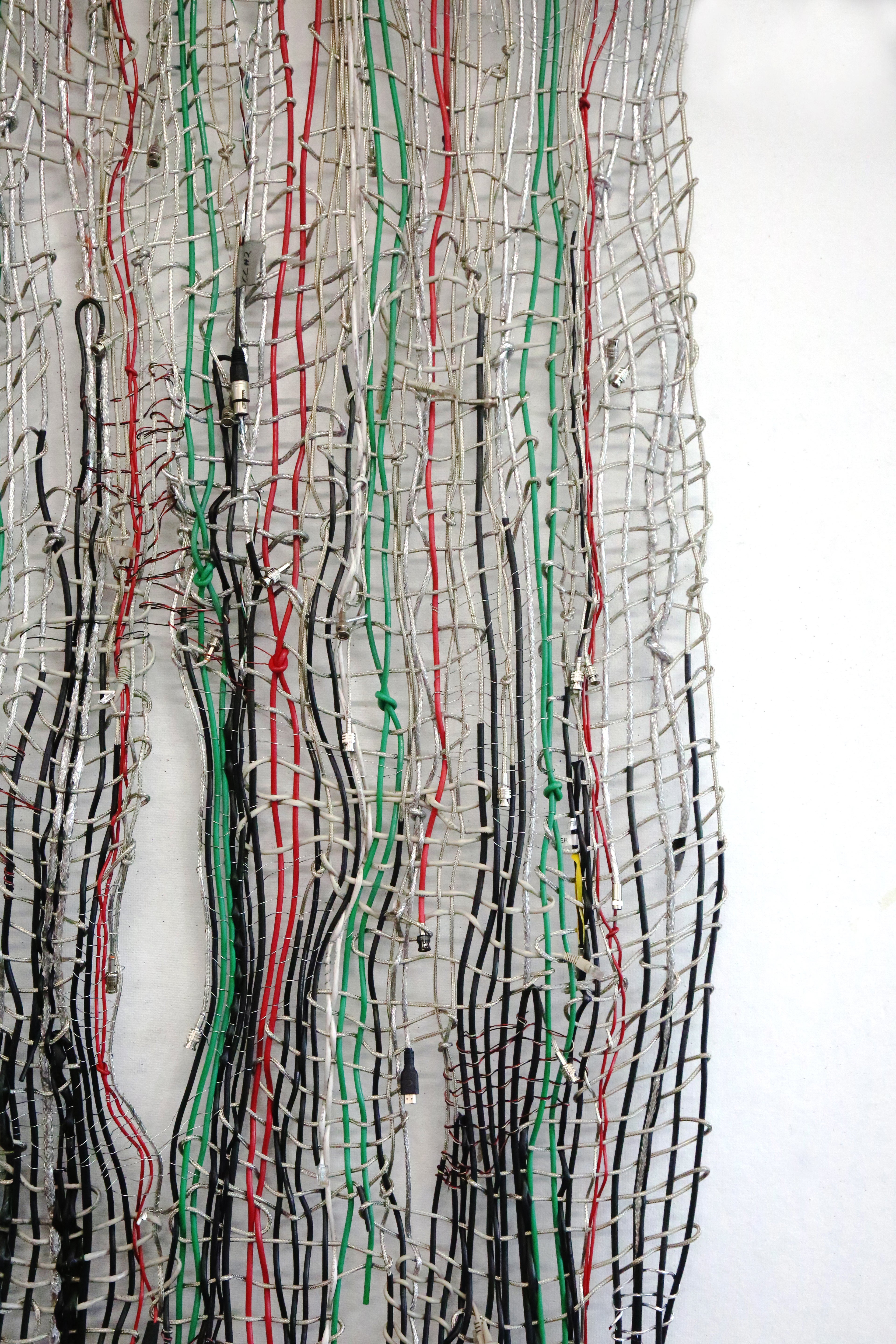 Vertical off center close up of weave made from various cables with the red, green, and black cables highlighted against the other stripped silver wires against a white background.