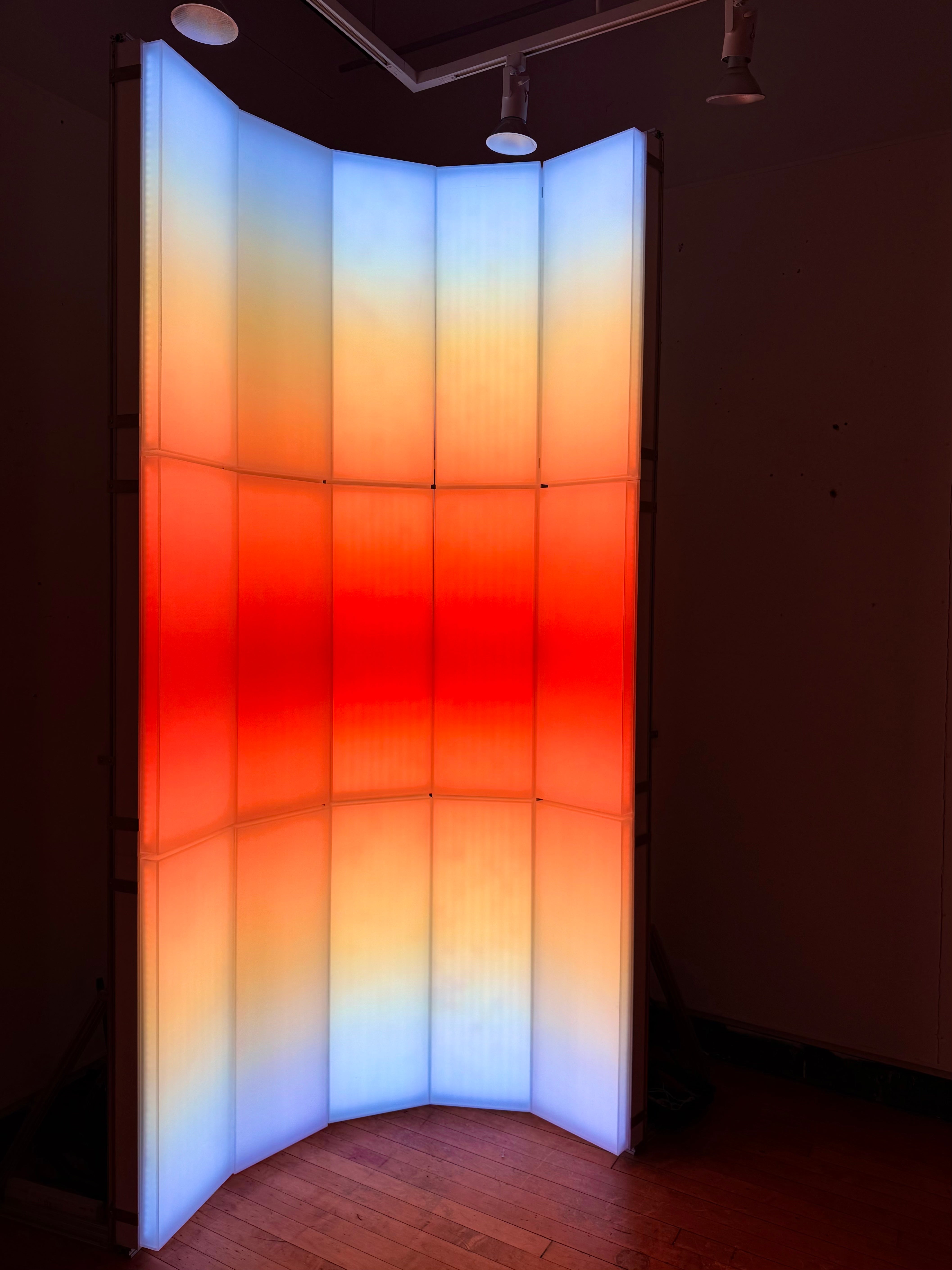 A light-based installation composed of vertical translucent panels, with blue, orange, and red gradients that evoke the color transition of sunrise and sunset.