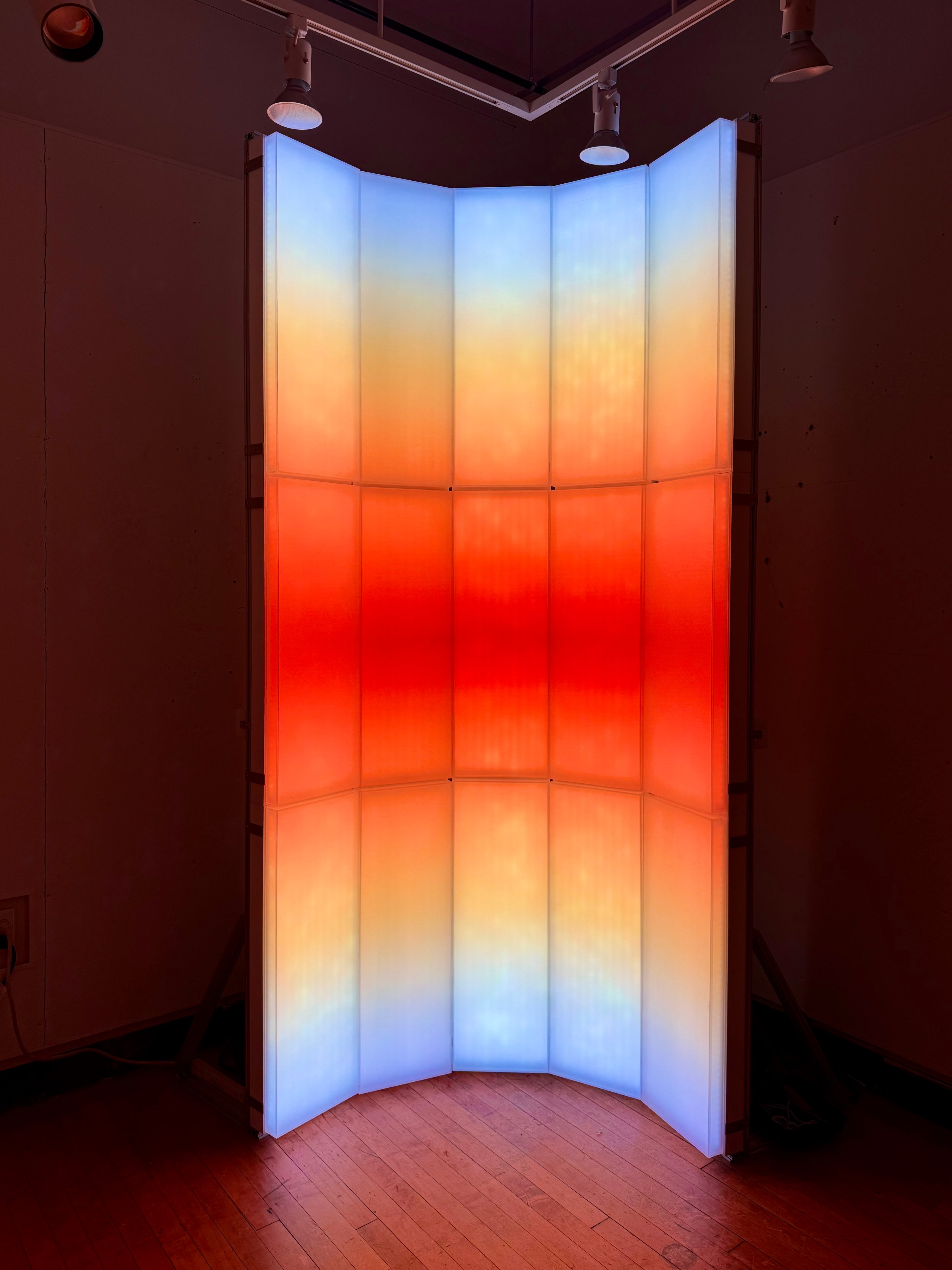 A light-based installation composed of vertical translucent panels, with blue, orange, and red gradients that evoke the color transition of sunrise and sunset.