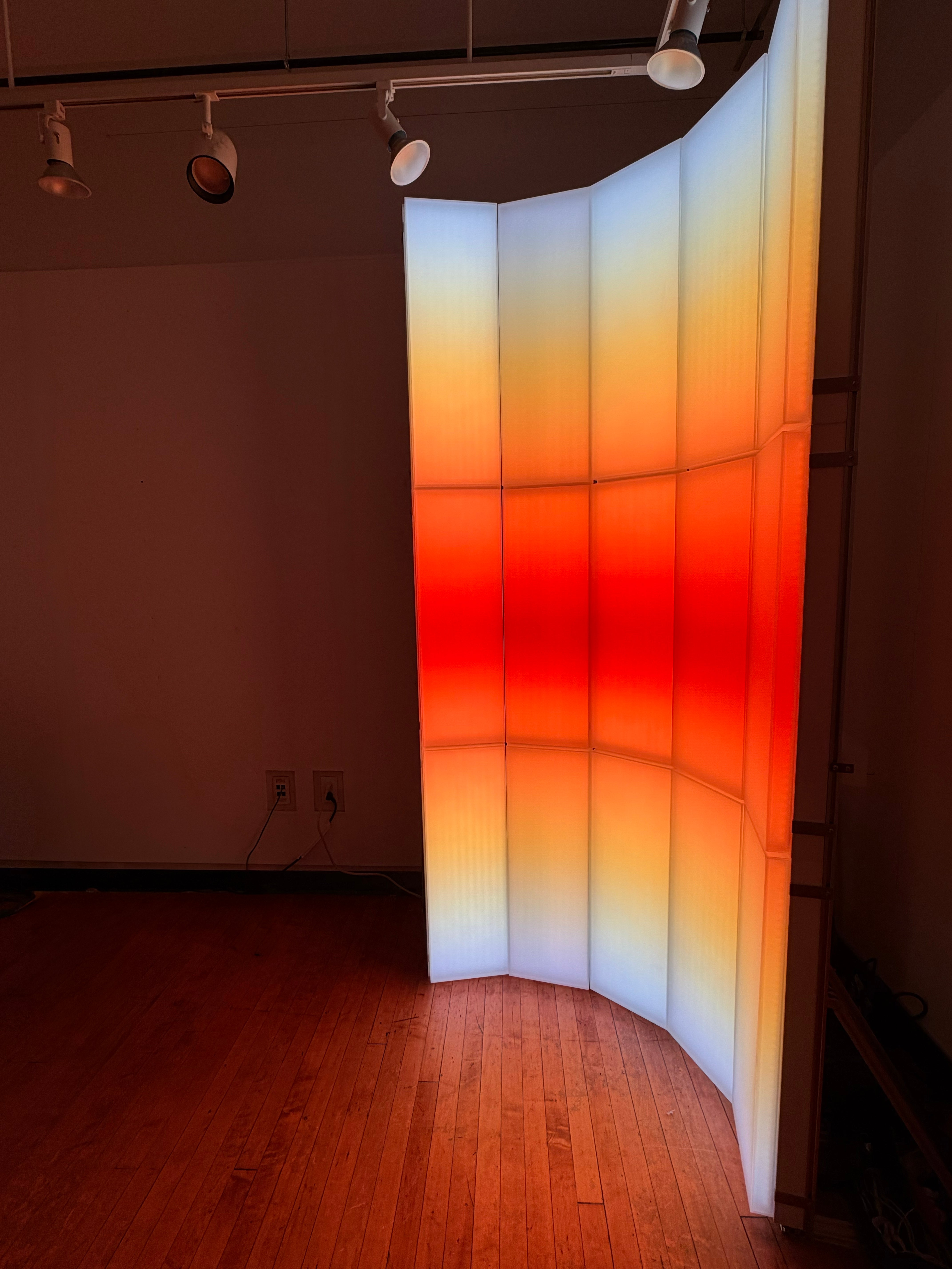 A light-based installation composed of vertical translucent panels, with blue, orange, and red gradients that evoke the color transition of sunrise and sunset.