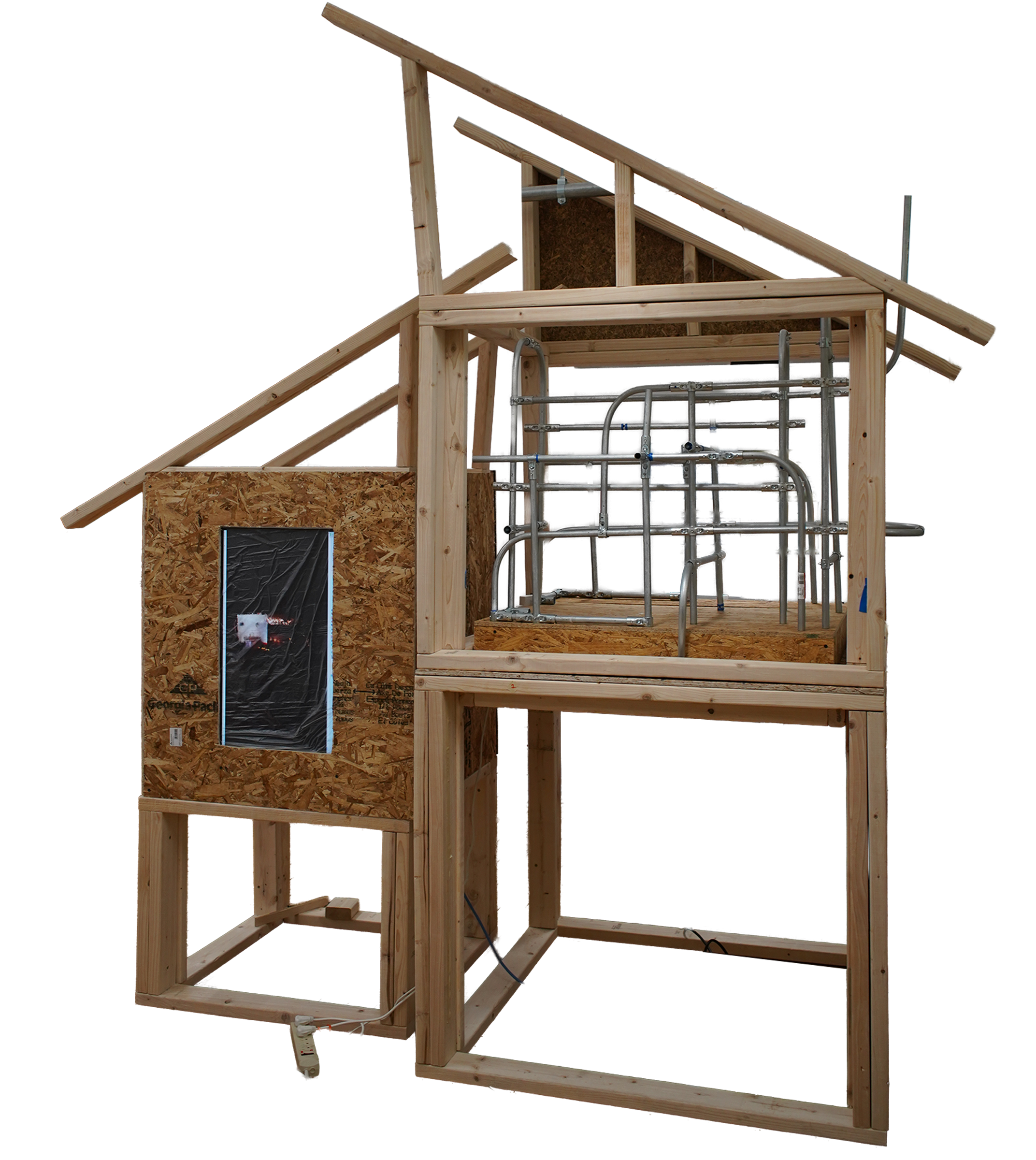 A multi-level architectural installation built from exposed raw lumber, resembling an unfinished house frame. The right side features an upper level densely filled with a geometric network of intersecting, bent metal conduits. 