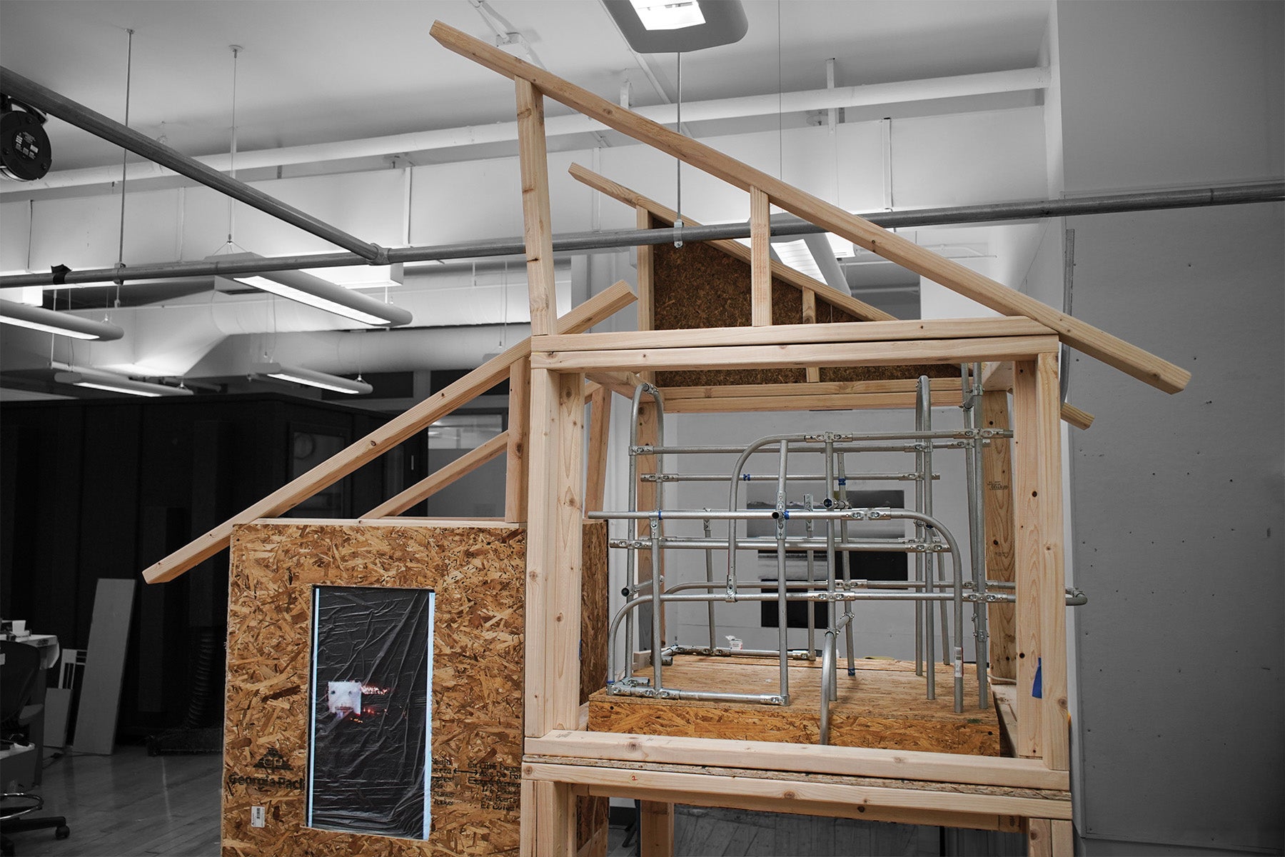 A close-up view of the architectural installation frame, constructed from raw lumber. On the left side, a section is enclosed by a textured particleboard (OSB) wall. 