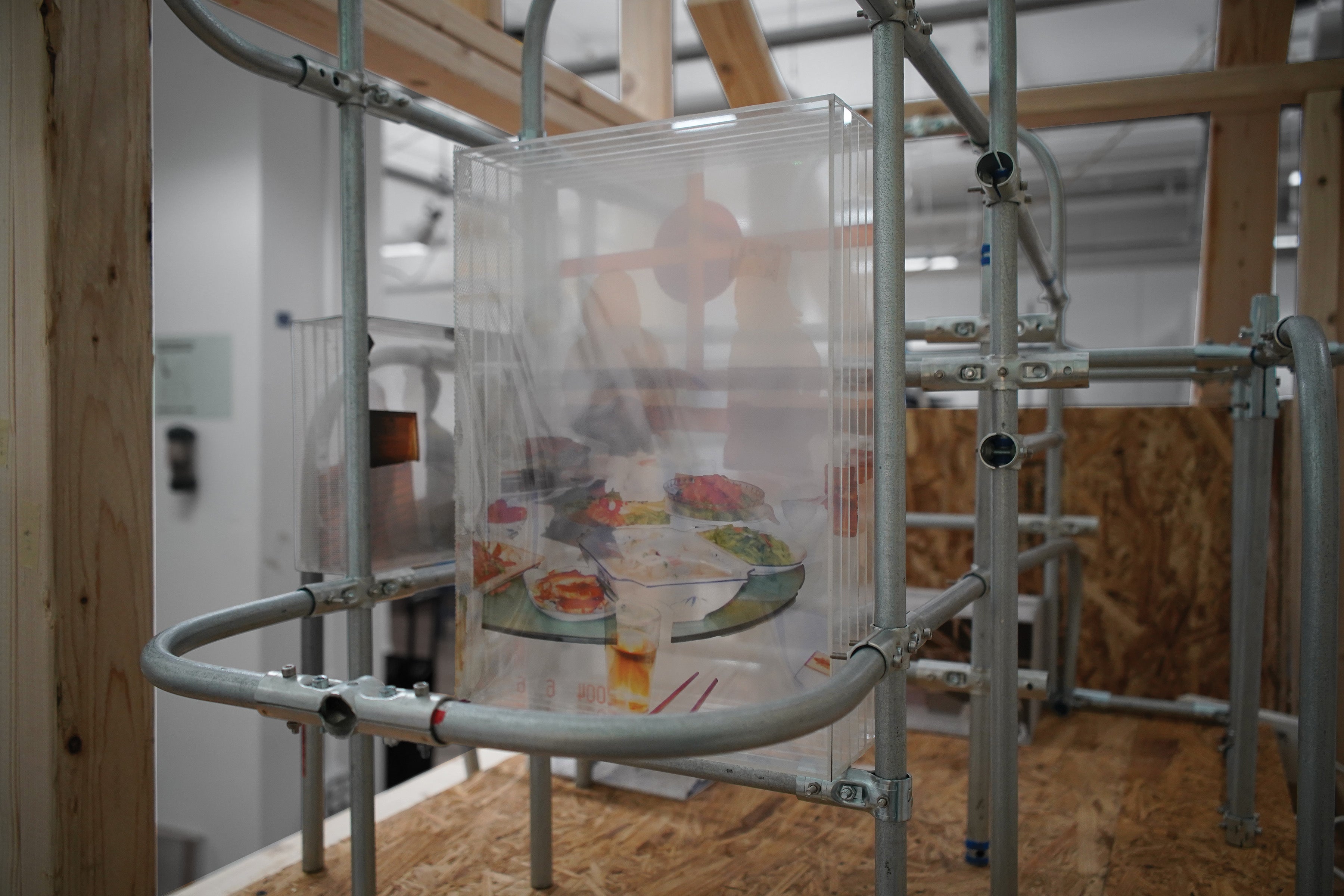 A detailed close-up of the art installation frame built from raw lumber and curved metal conduits. A multi-layered transparent acrylic box, held within the structure, contains a deconstructed archival photograph of a dinner table scene. 