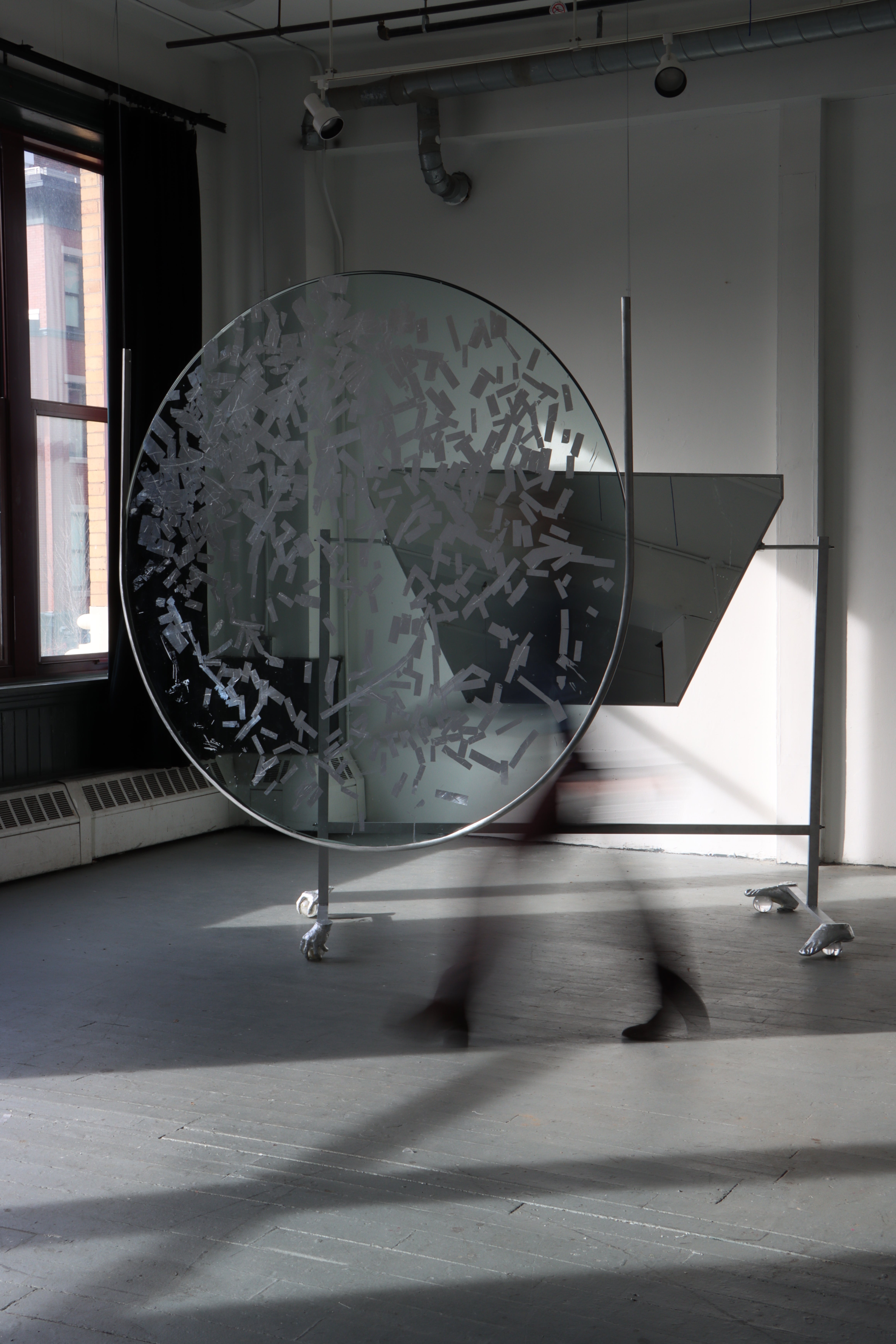  4ft diameter piece of glass hands supsended in a room, with part of its surfaced gilded silver. A person in motion passes behind it. Behind them is large piece of mirror angled on a aluminum stand, with feet made of hands and feet on crystal balls. 