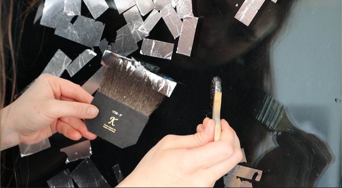 A close up shot of a person applying aluminum leaf to a glass with a brush for the leaf in one hand, and a brush for size in the other. The glass is against a black  background and the veins and wrinkles of the metal contrast with the smoothness of the glass surface.