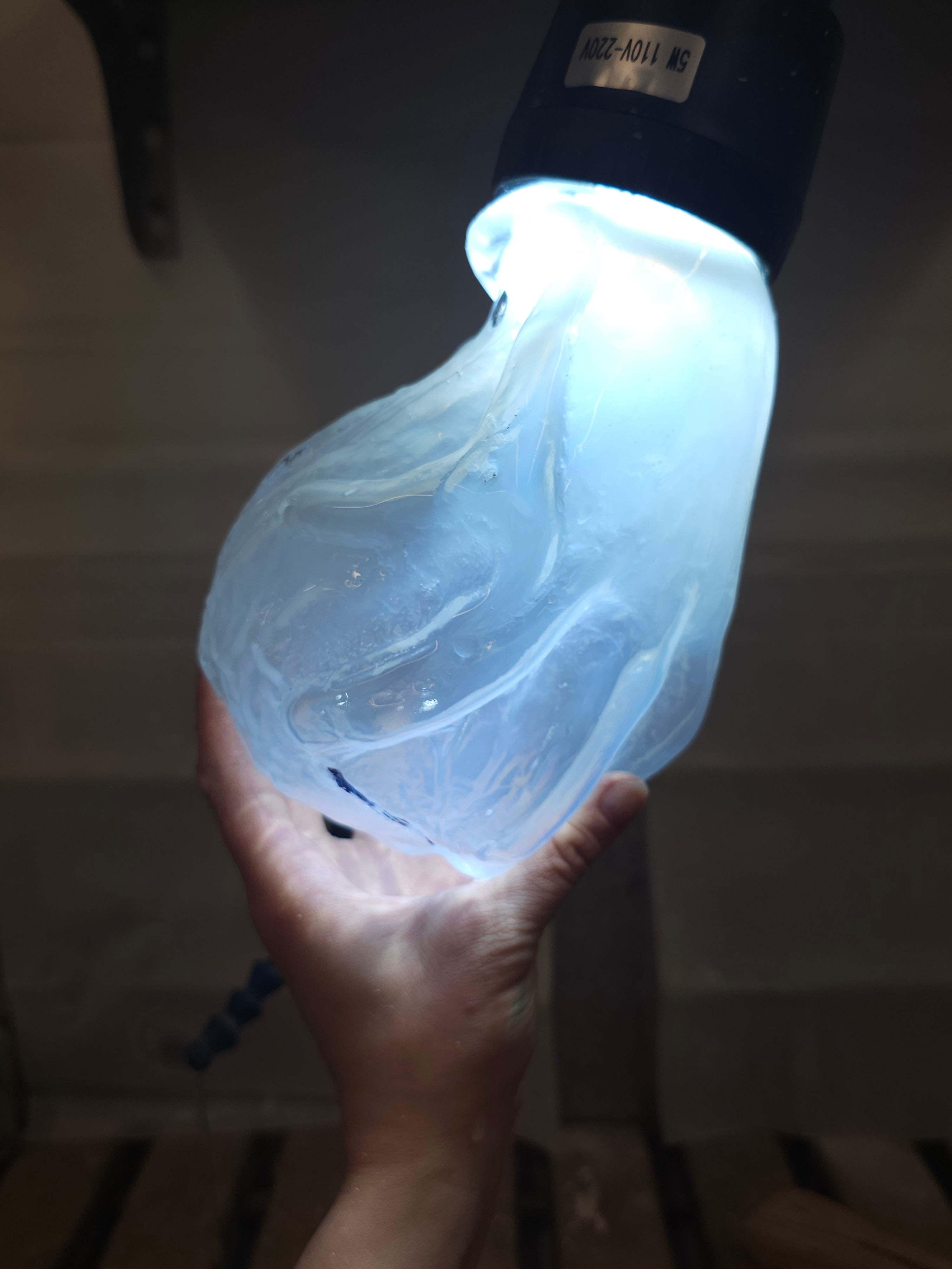 Against a darkened background, a pale hand holes up a piece of blown glass the shape of a hand grasping a sphere-ish shape, glowing blue from within. 